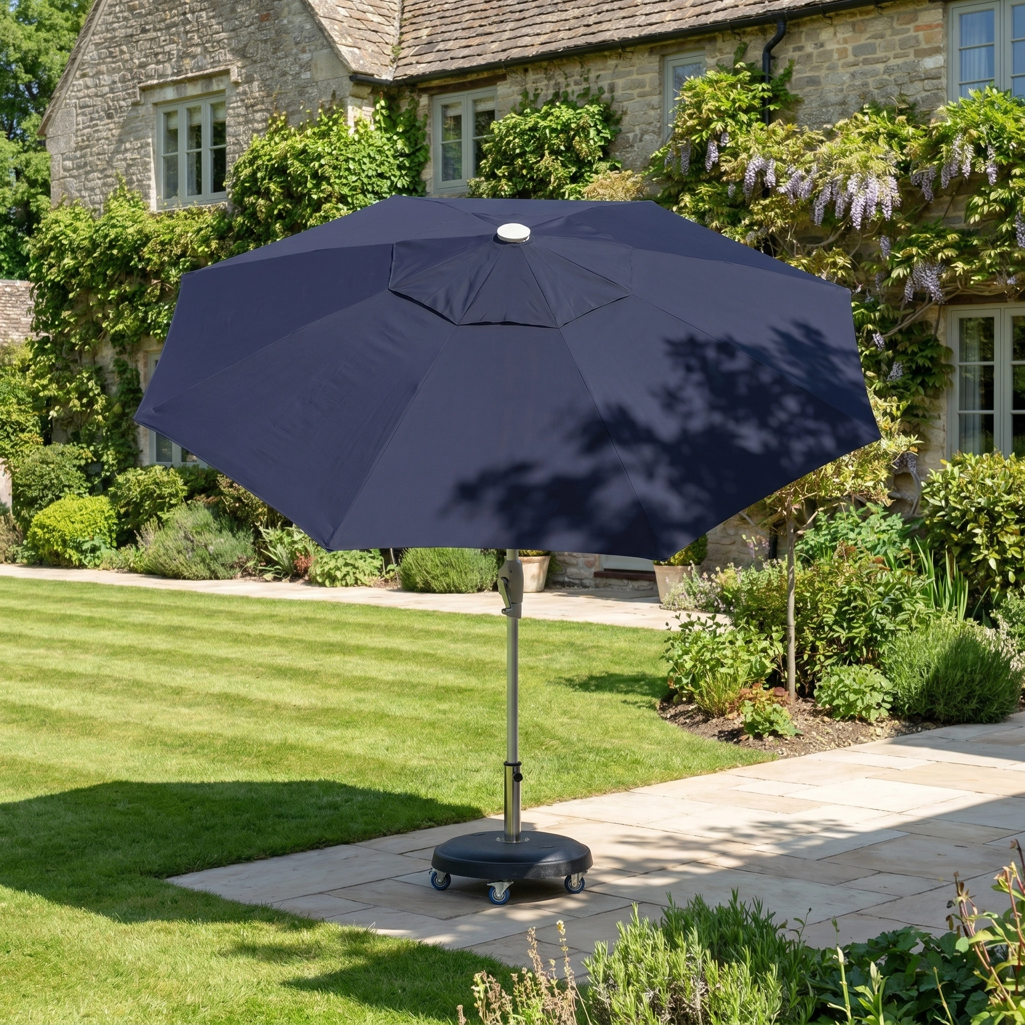 The Zelos 2.5M Round Parasol with LED Lighting in Navy stands on a stone terrace in a sunny, green garden beside a house.