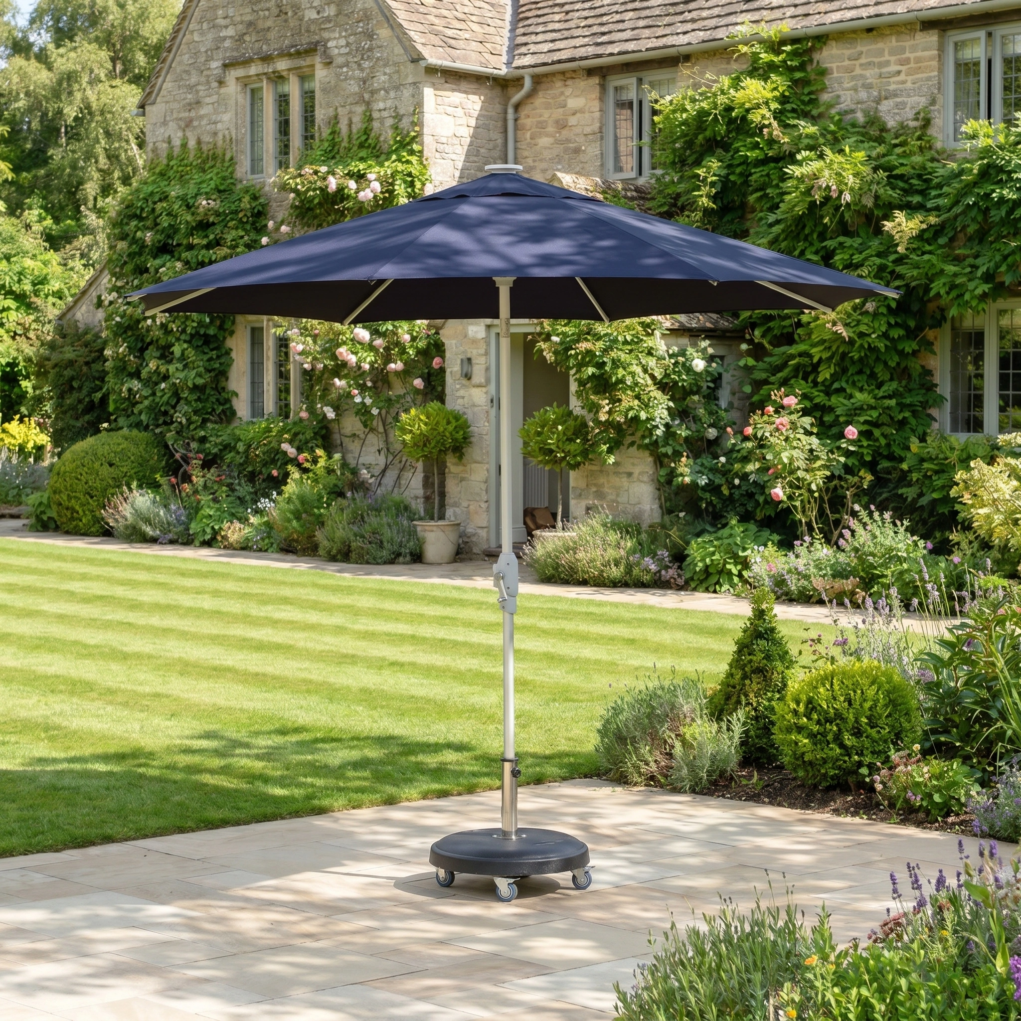 The Zelos 2.5M Round Parasol with LED Lighting in Navy stands on a stone terrace in a garden beside a stone house covered in greenery.