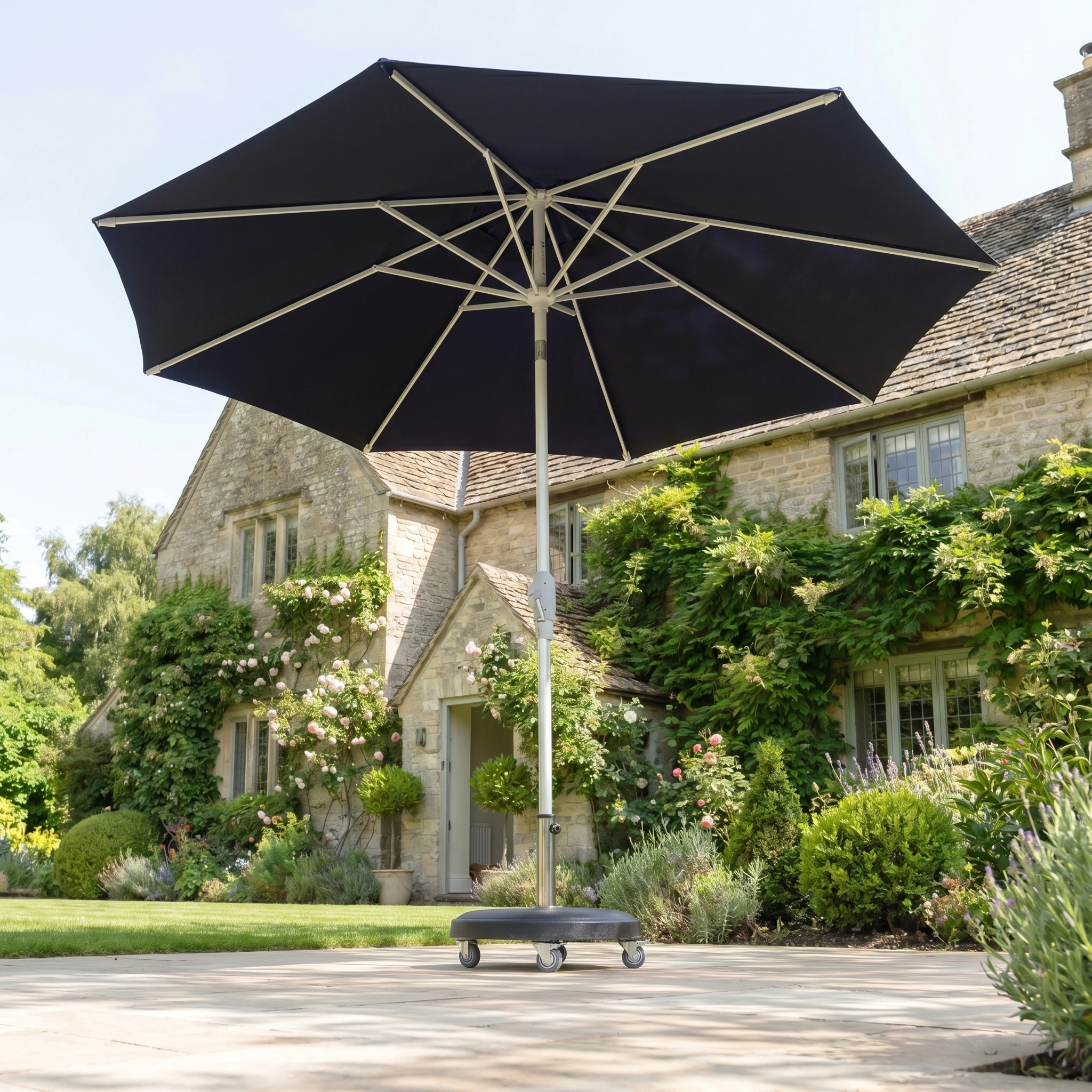A Zelos 2.5M Round Parasol with LED Lighting in navy stands open on wheels in front of a stone house with a lush garden.