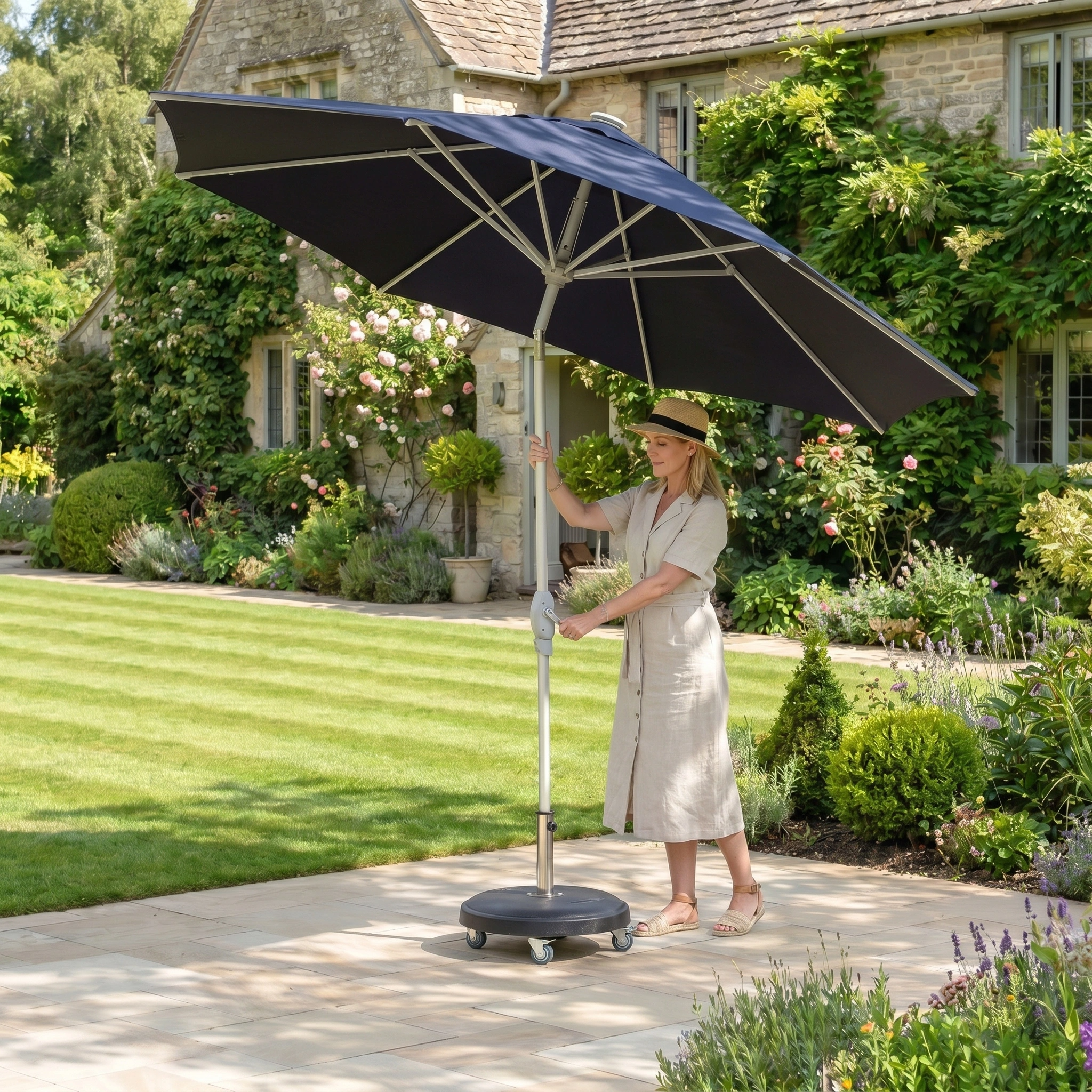 A woman in a light dress adjusts the Zelos 2.5M Round Parasol with LED Lighting in Navy in her sunny garden beside a stone house, bringing style and comfort to her outdoor space.