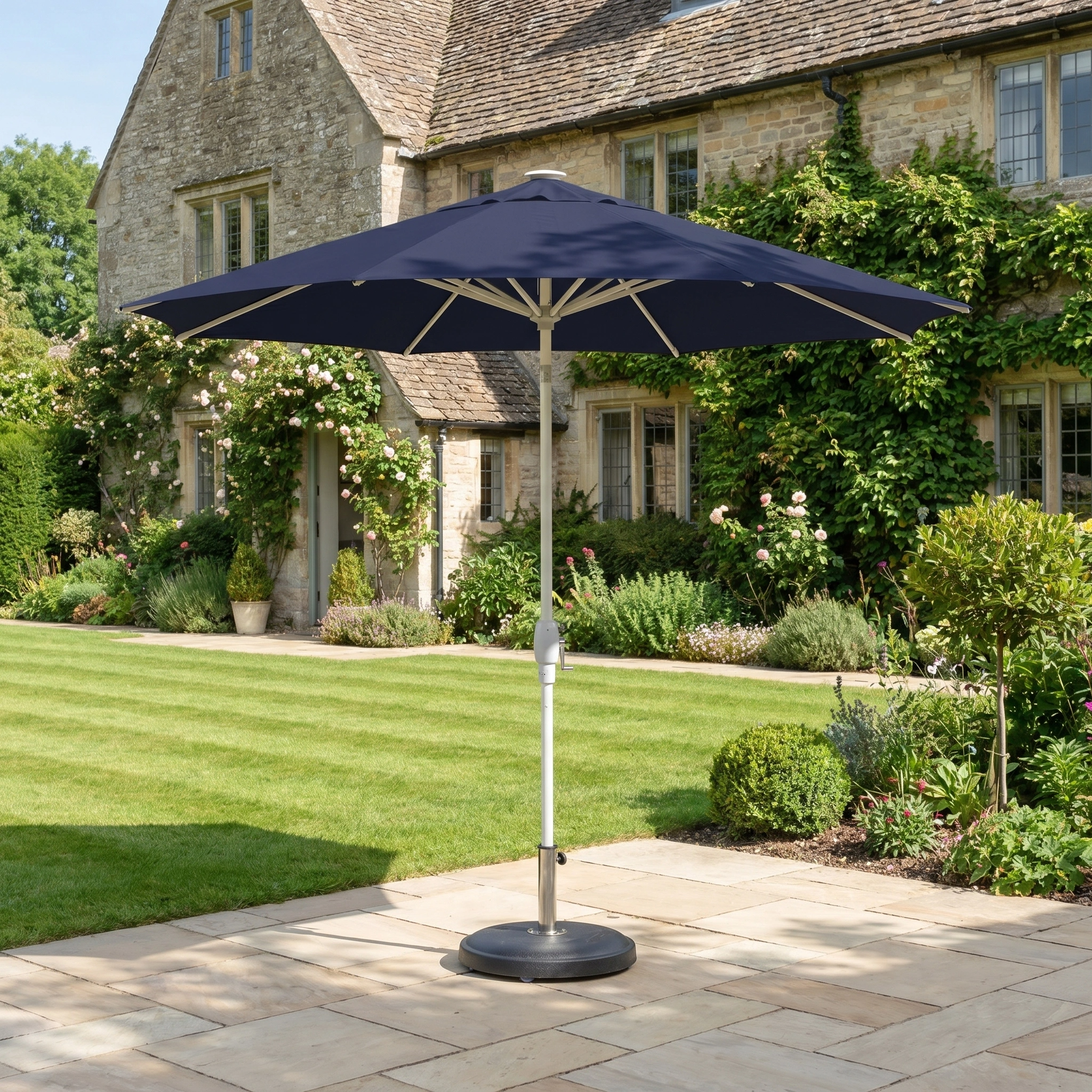 The Zelos 2.5M Round Parasol with LED Lighting in Navy stands on a stone terrace in a garden, with a large stone house visible in the background.