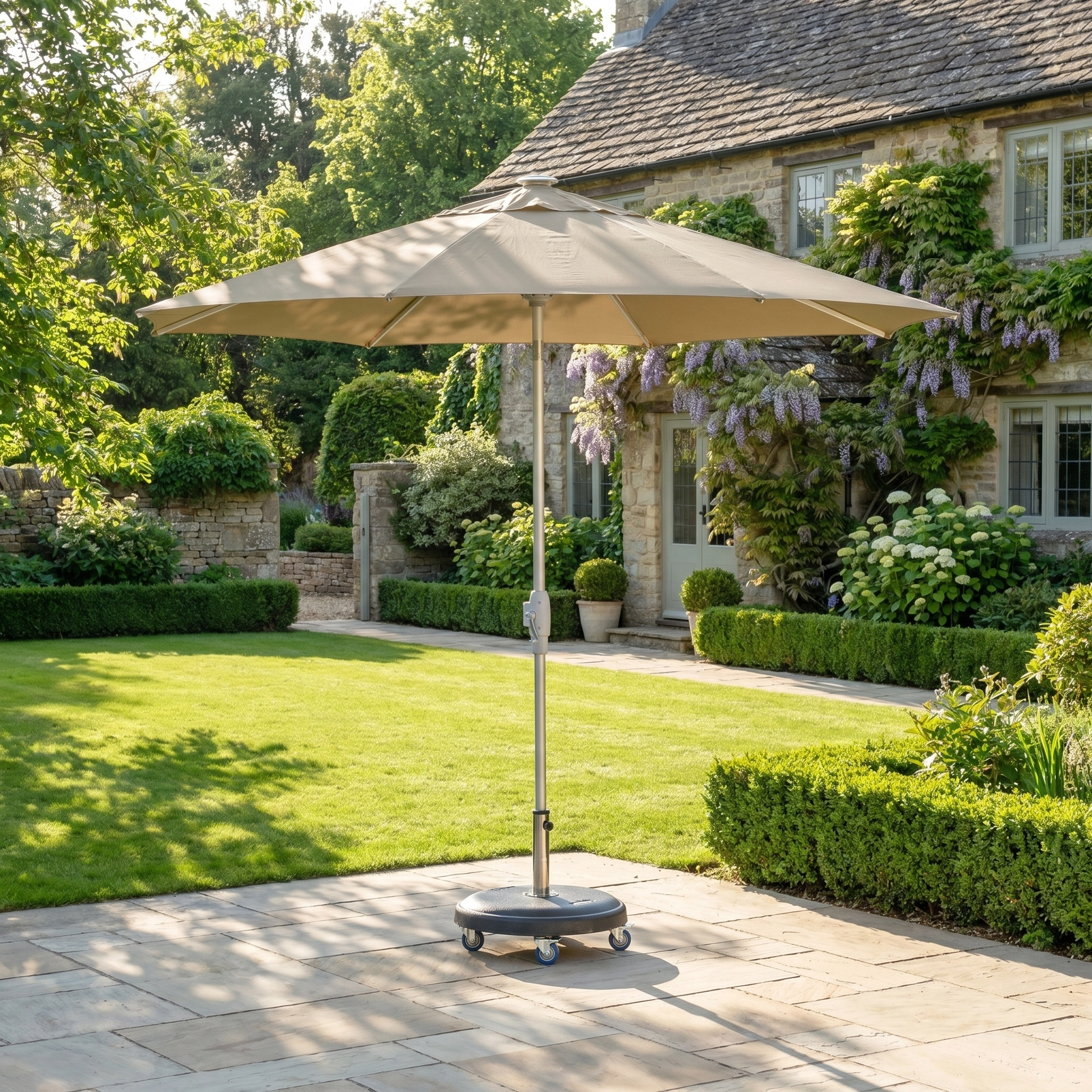 The Zelos 2.5M Round Parasol with LED Lighting in Taupe stands on a stone patio in a lush garden, providing UV protection and stylish shade with views of the stone house beyond.