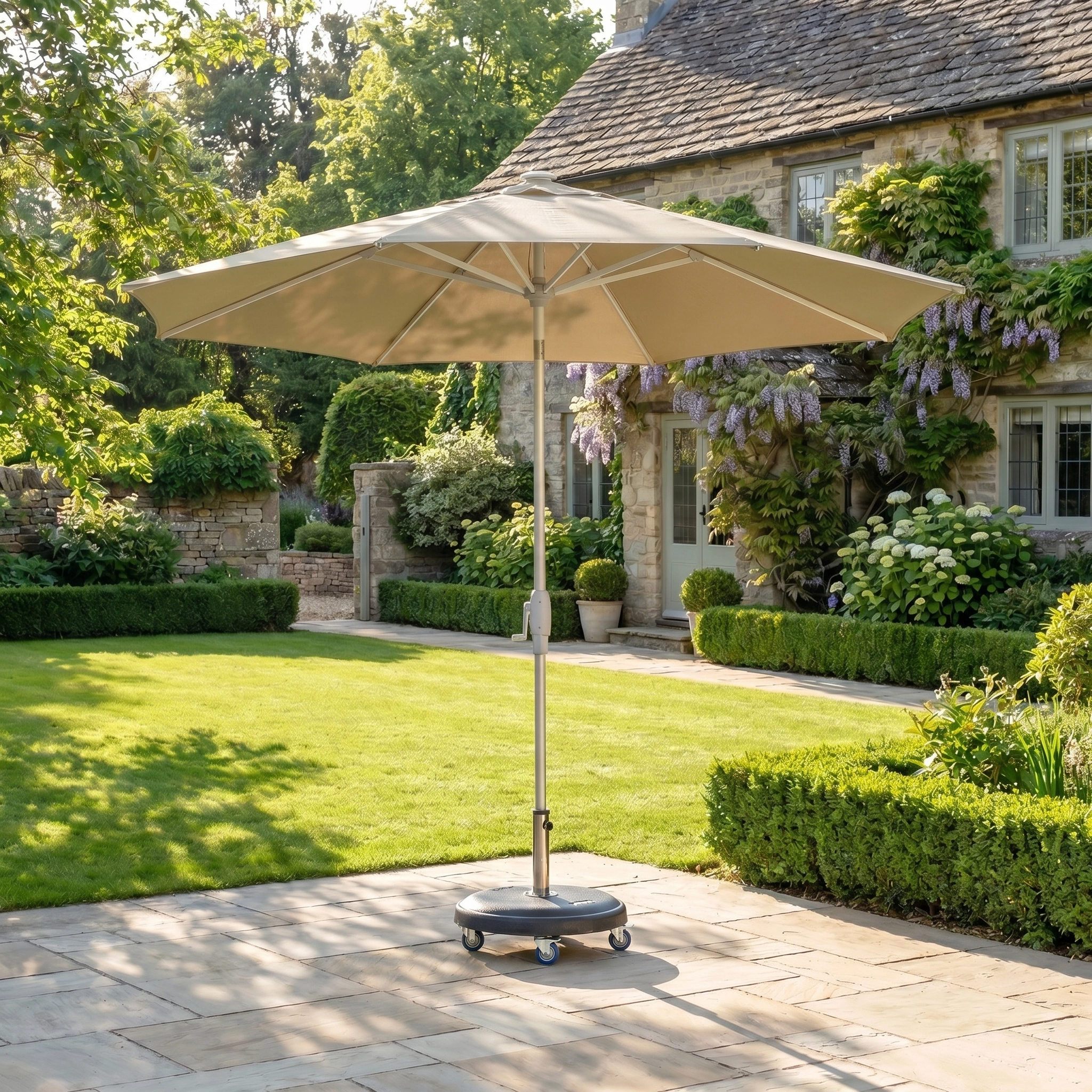 A Zelos 2.5M Round Parasol with LED Lighting in taupe stands on a stone terrace in a lush garden beside a cottage, offering stylish shade and UV protection.