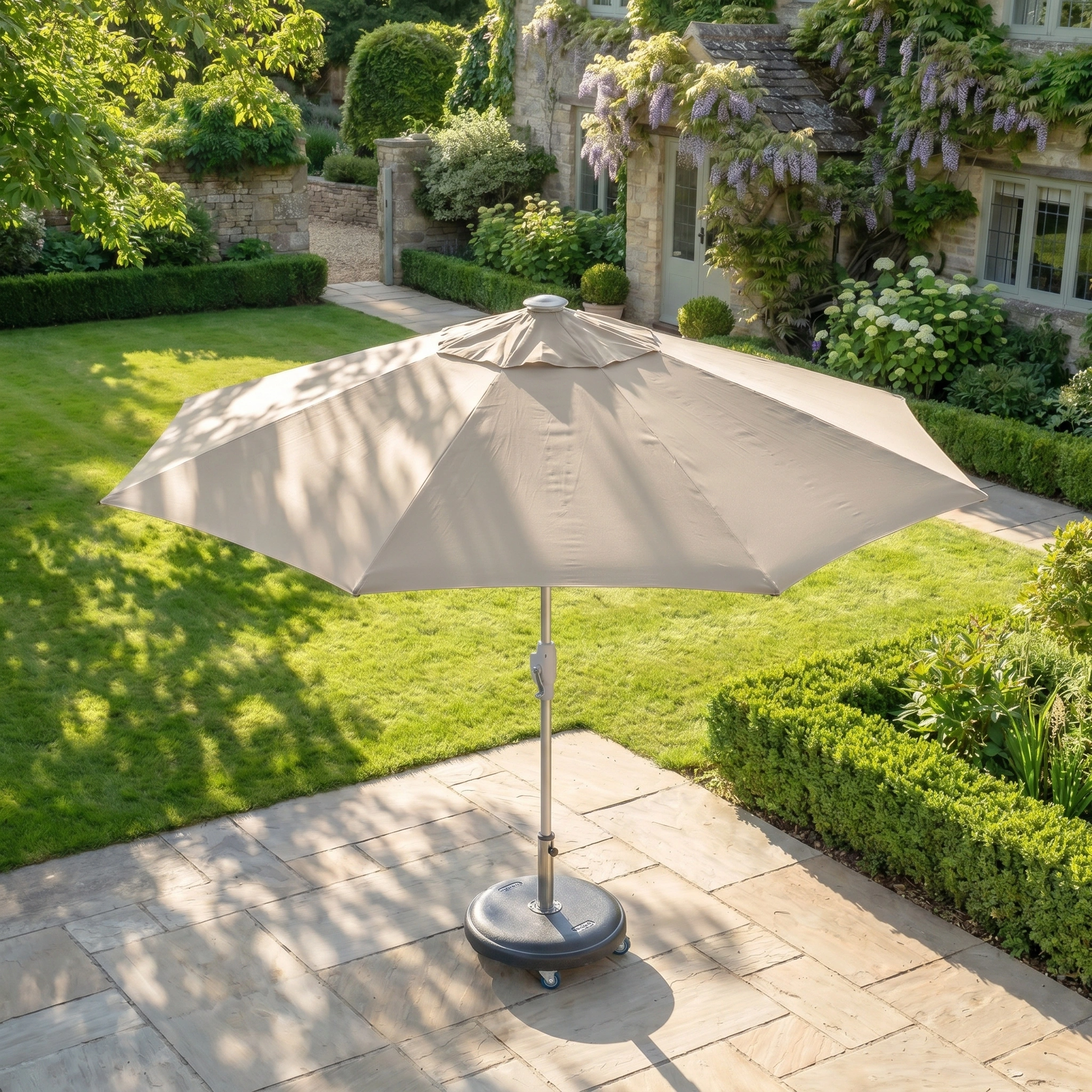 The Zelos 2.5M Round Parasol with LED Lighting in Taupe provides shade and UV protection on a stone patio in a lush garden, with a stone house in the background.
