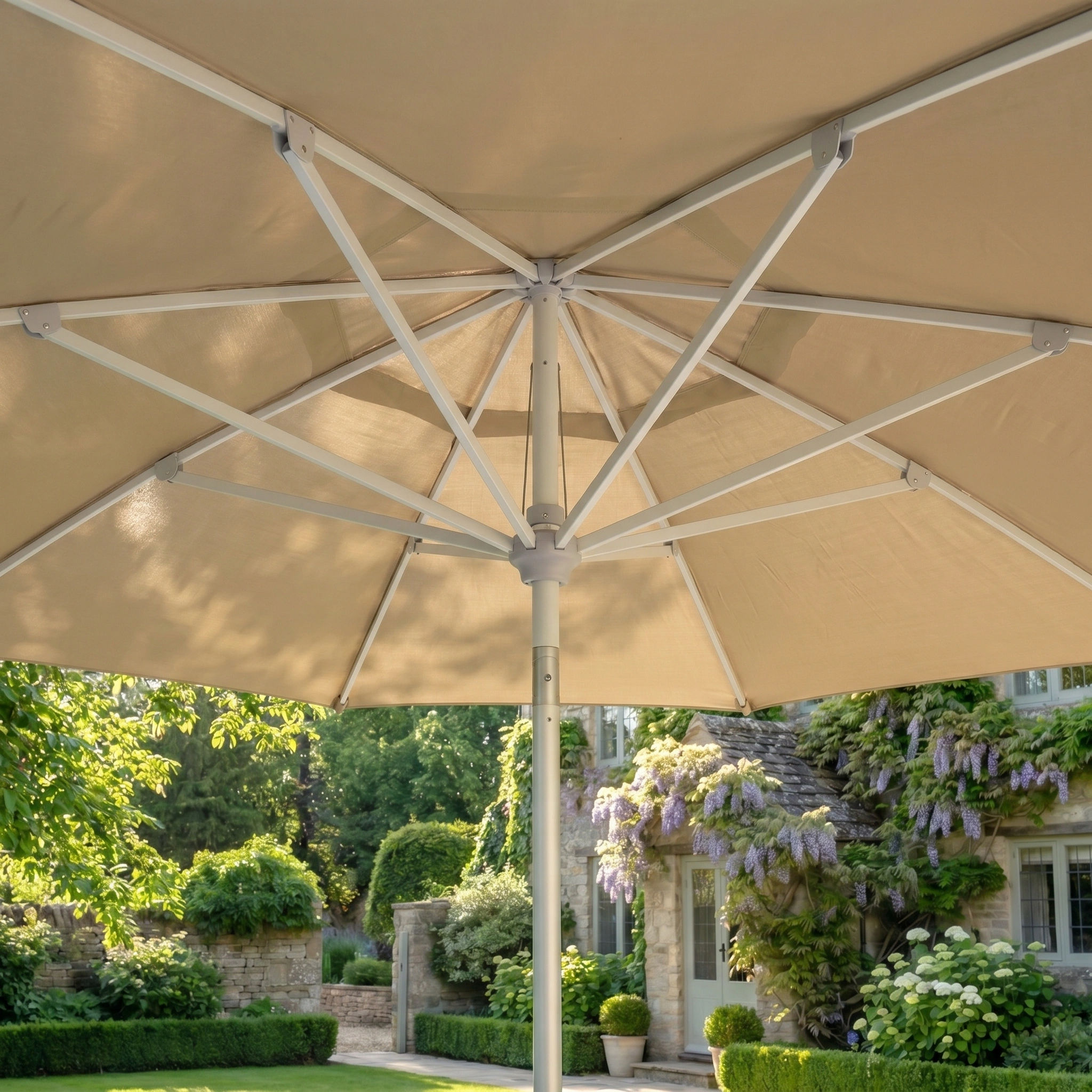 Enjoy the view from beneath the Zelos 2.5M Round Parasol with LED Lighting in Taupe, set in a lush garden beside a stone house and blooming wisteria.