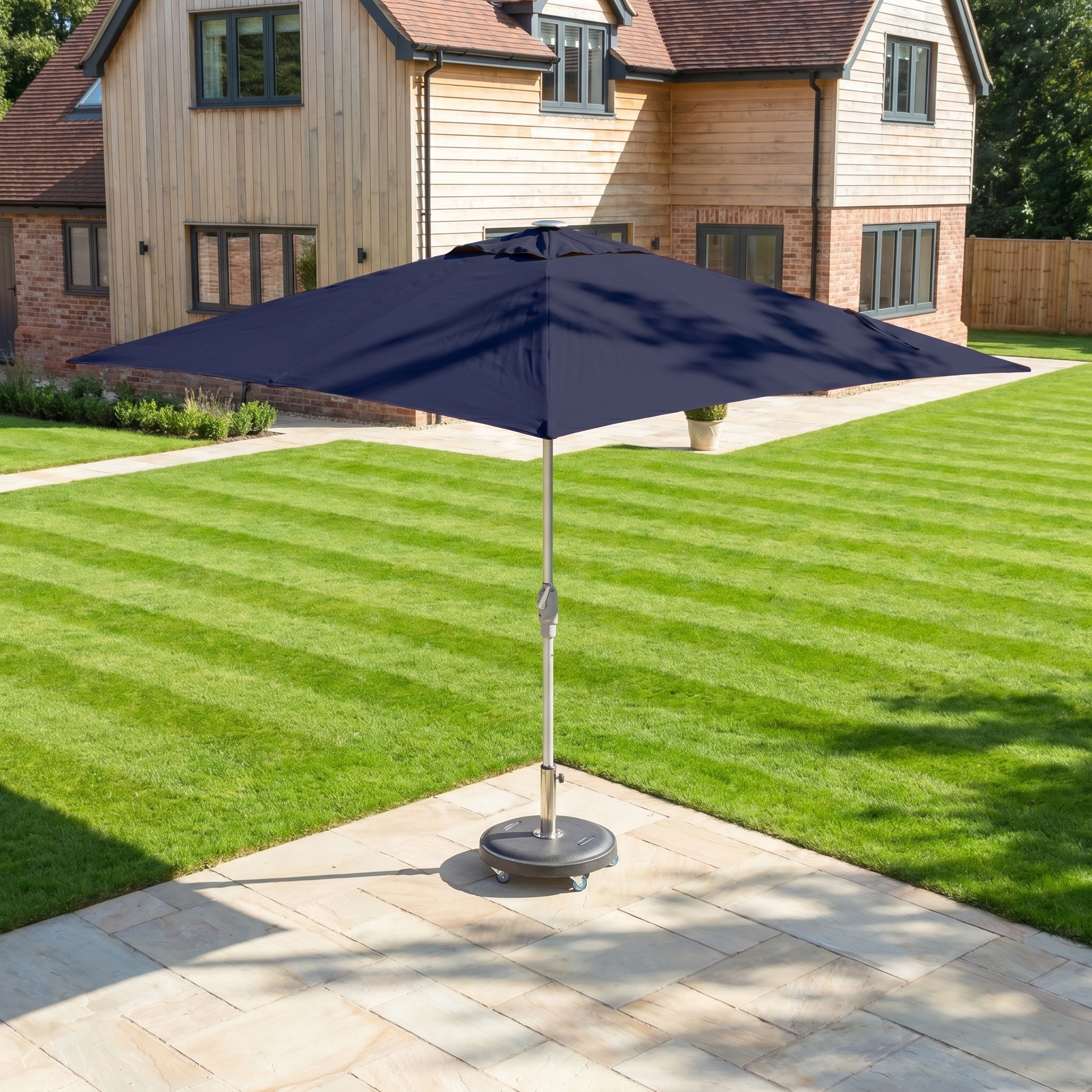 The Zelos 2.5m Square Parasol with LED Lighting in Navy offers UV protection and stands on a stone patio in a neatly mowed backyard by a modern house.