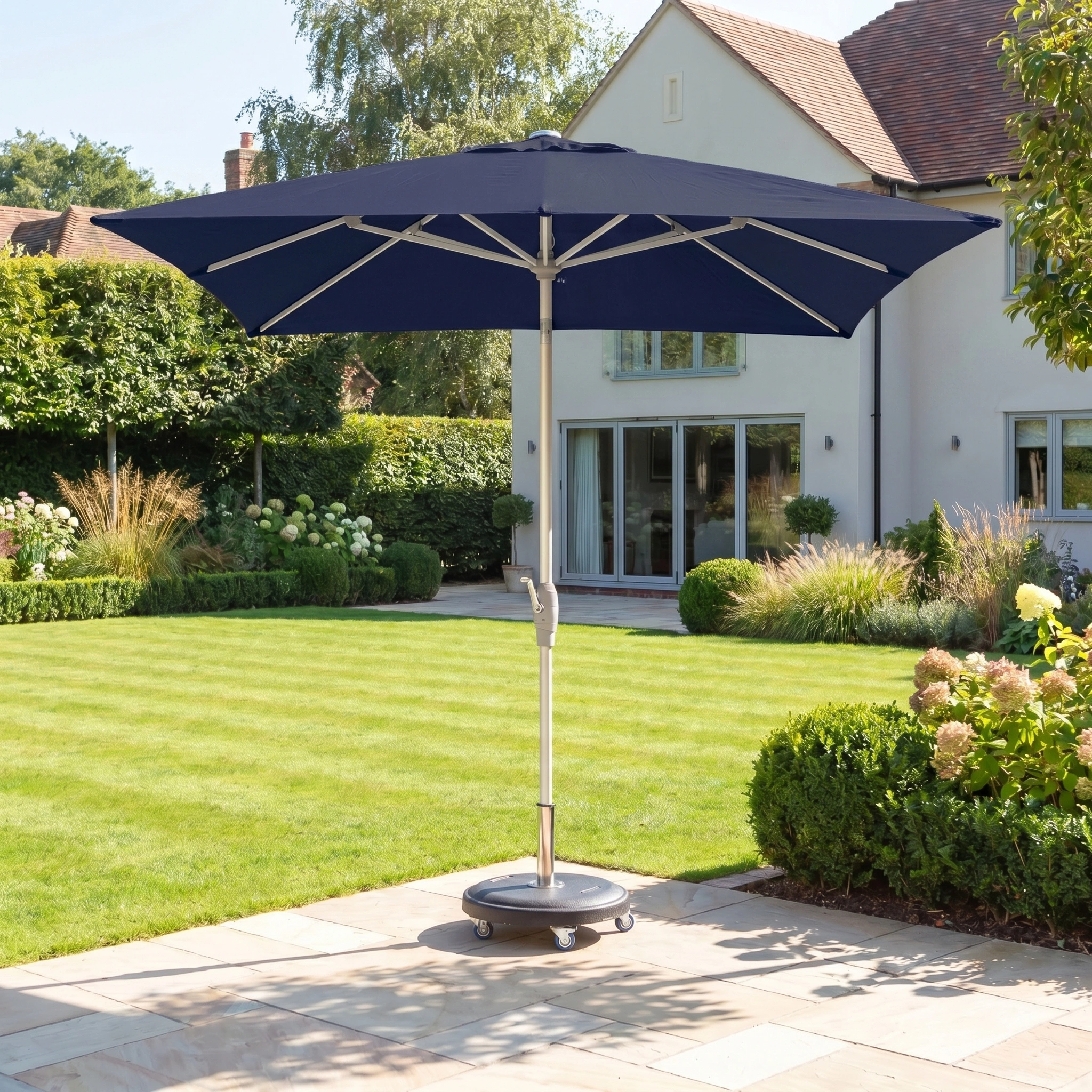 The Zelos 2.5m Square Parasol with LED Lighting in navy stands open on a sunny stone patio in a landscaped garden, providing stylish shade and UV protection.