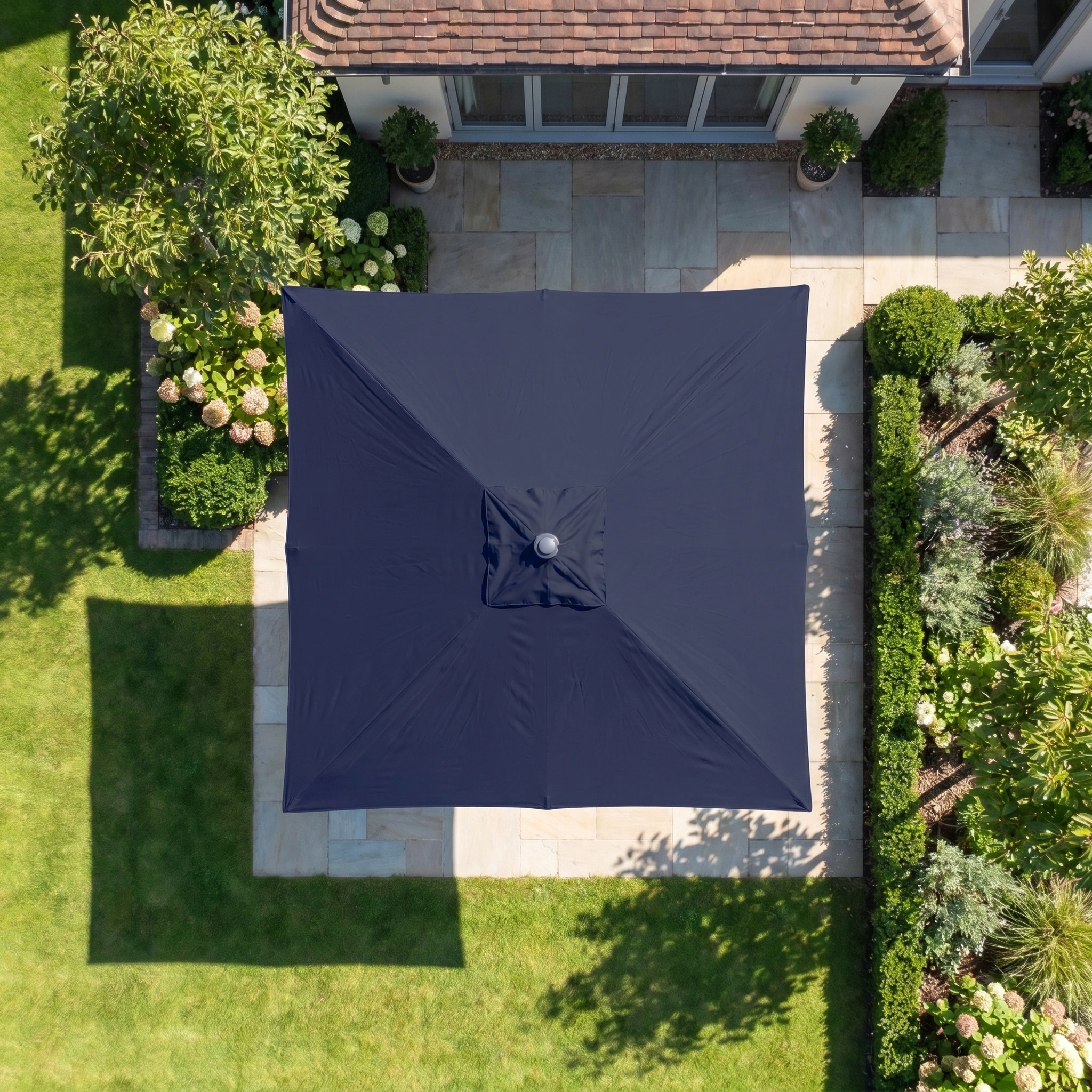 The Zelos 2.5m Square Parasol with LED Lighting in Navy provides UV-protected shade to a stone patio in a landscaped backyard, as seen from above.