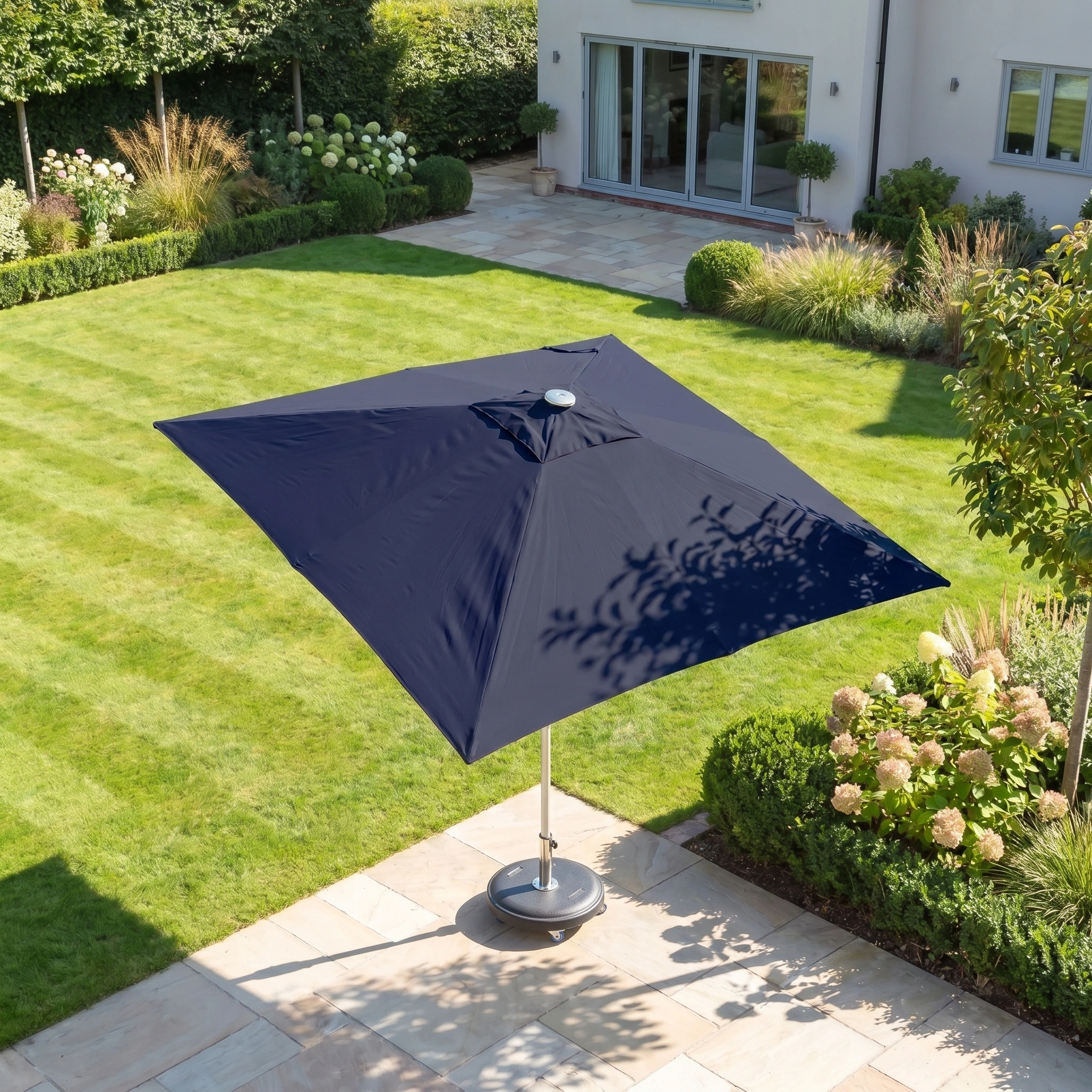 The Zelos 2.5m Square Parasol with LED Lighting in Navy stands on a sunny stone terrace beside a modern house, offering stylish UV protection in your landscaped garden.