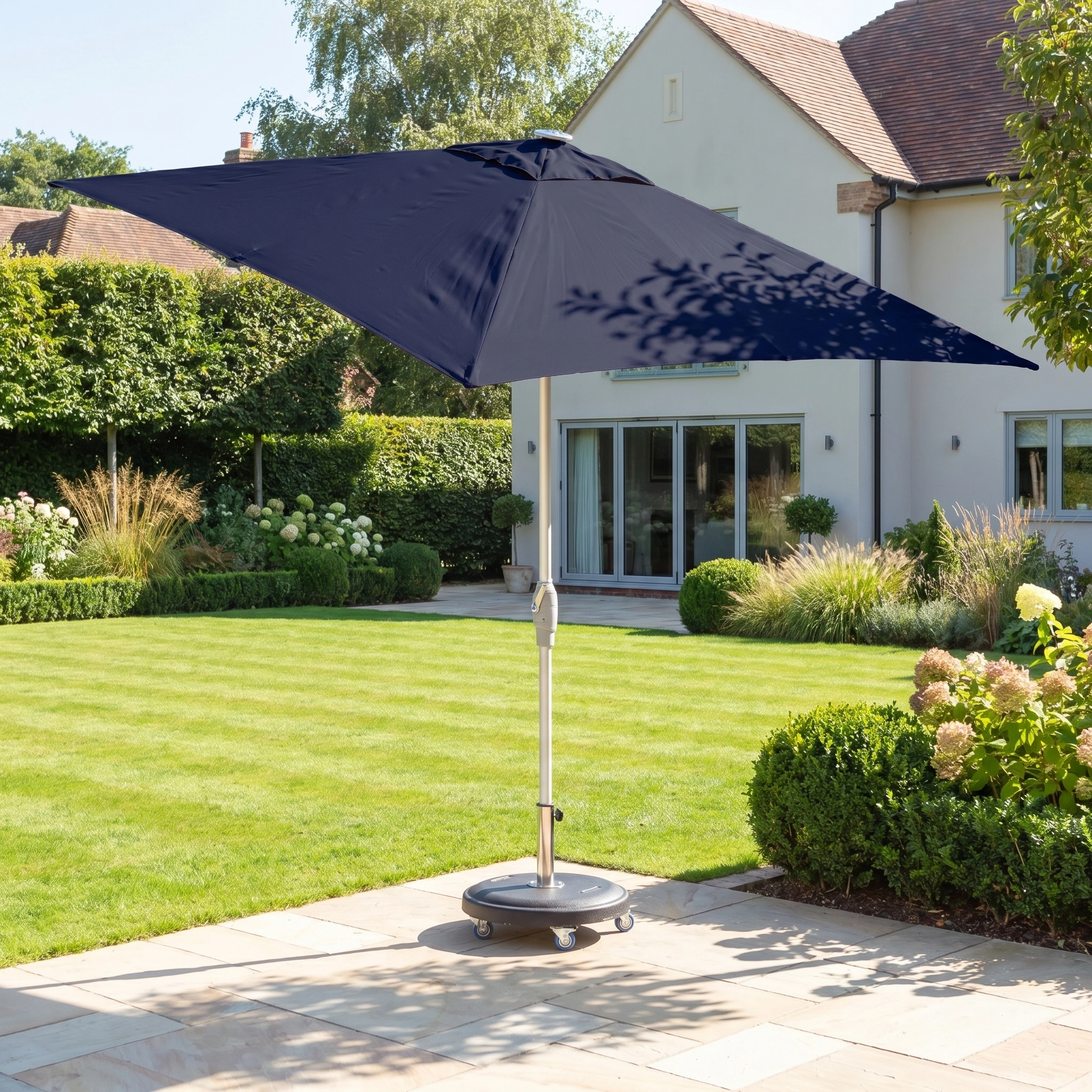 The Zelos 2.5m Square Parasol with LED Lighting in Navy stands open on a stone terrace in a landscaped backyard garden.