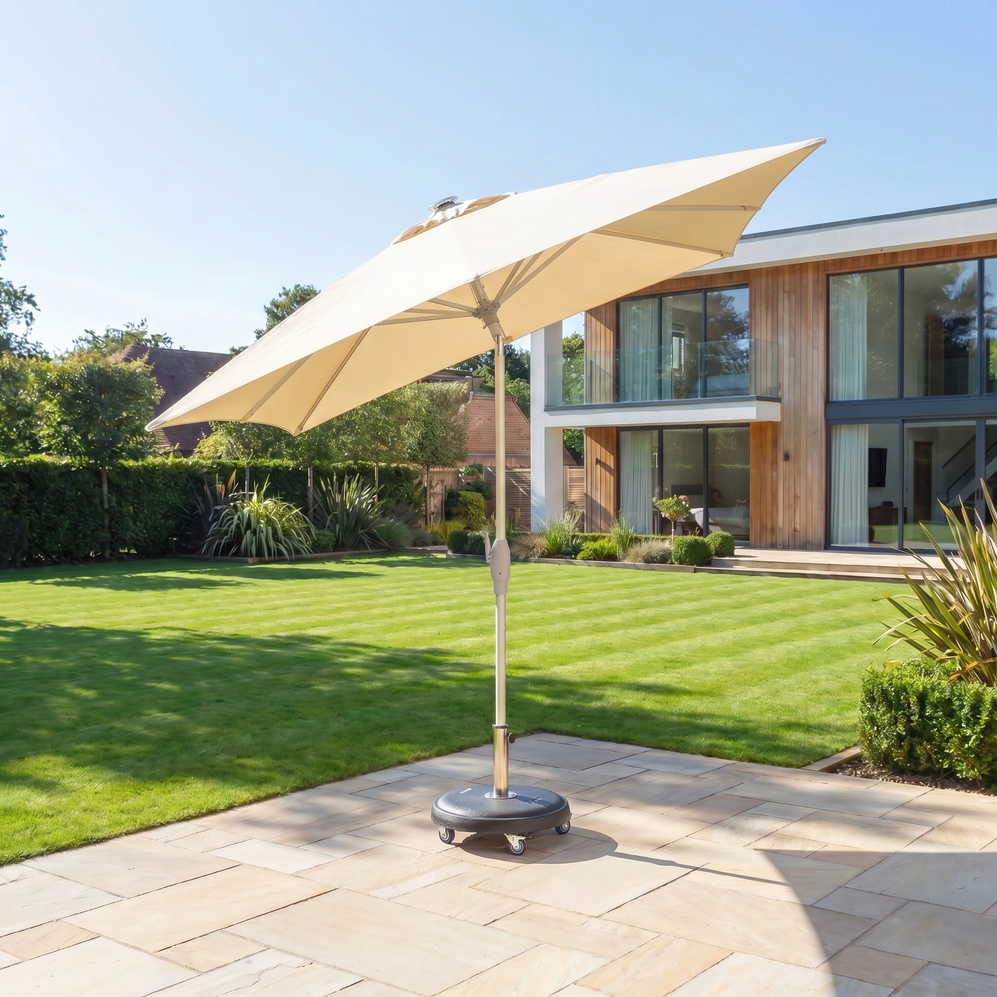 The Zelos 2.5m Square Parasol with LED Lighting in Cream is displayed on a stone terrace in a modern garden with a lawn and a contemporary house in the background.