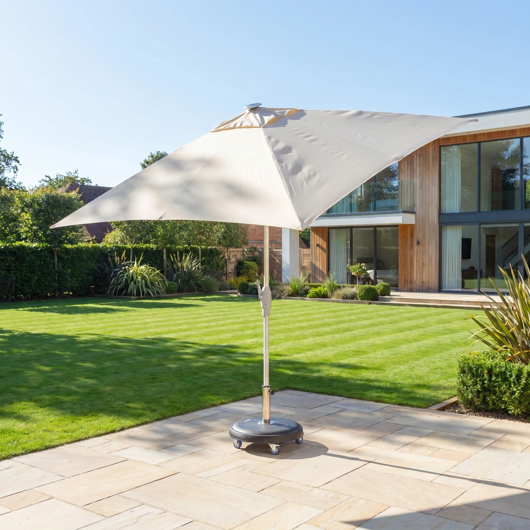The Zelos 2.5m Square Parasol with LED Lighting in Cream, set on a wheeled base, provides excellent UV protection for your patio against a backdrop of manicured lawn and modern house.