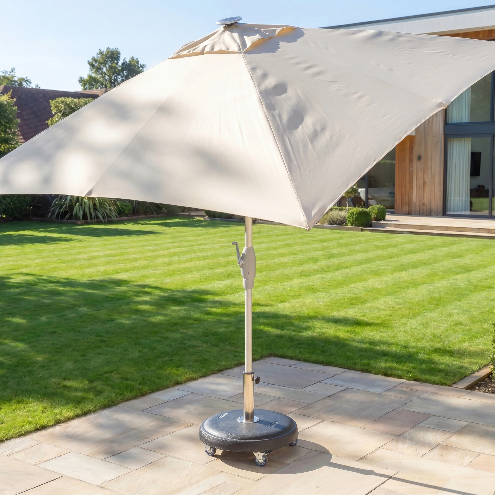 The Zelos 2.5m Square Parasol with LED Lighting in Cream stands on a stone patio, set against a green lawn and a modern house backdrop.