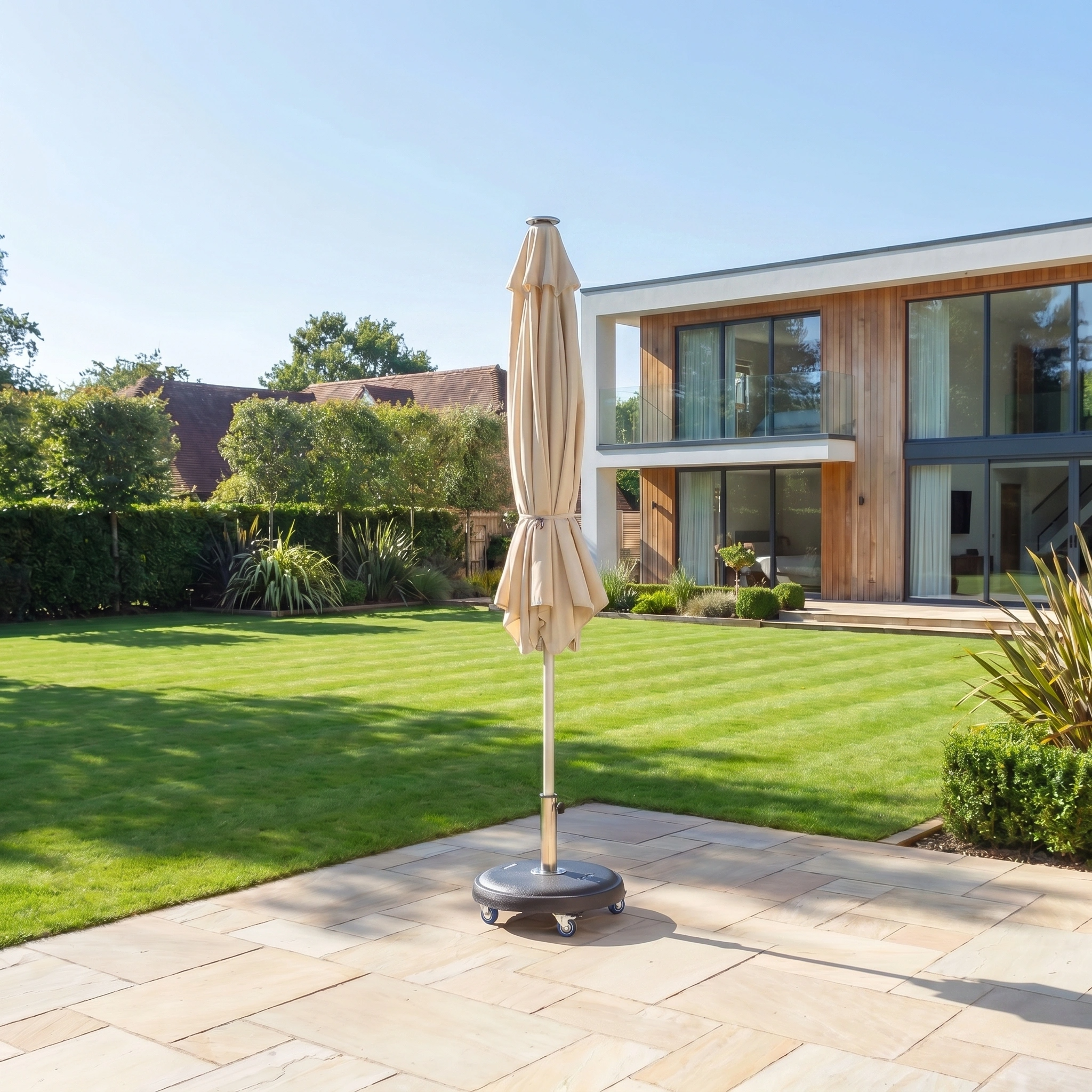 The Zelos 2.5m Square Parasol with LED Lighting in Cream stands closed on a stone terrace in a modern house garden, surrounded by a green lawn and shrubs.