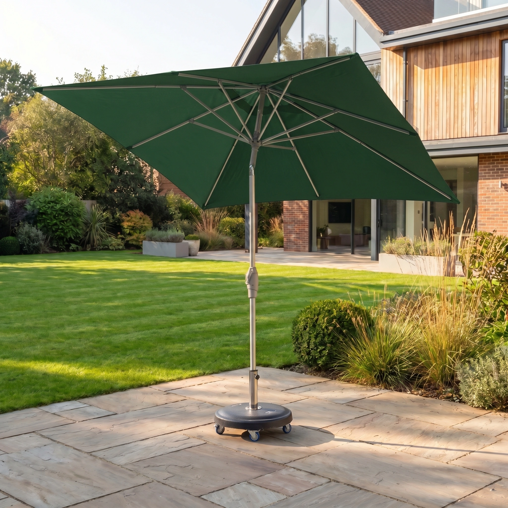 The Zelos 2.5m Square Parasol with LED Lighting in Green stands on a stone terrace in a modern garden, offering stylish shade and UV protection with the added feature of integrated LED lighting.