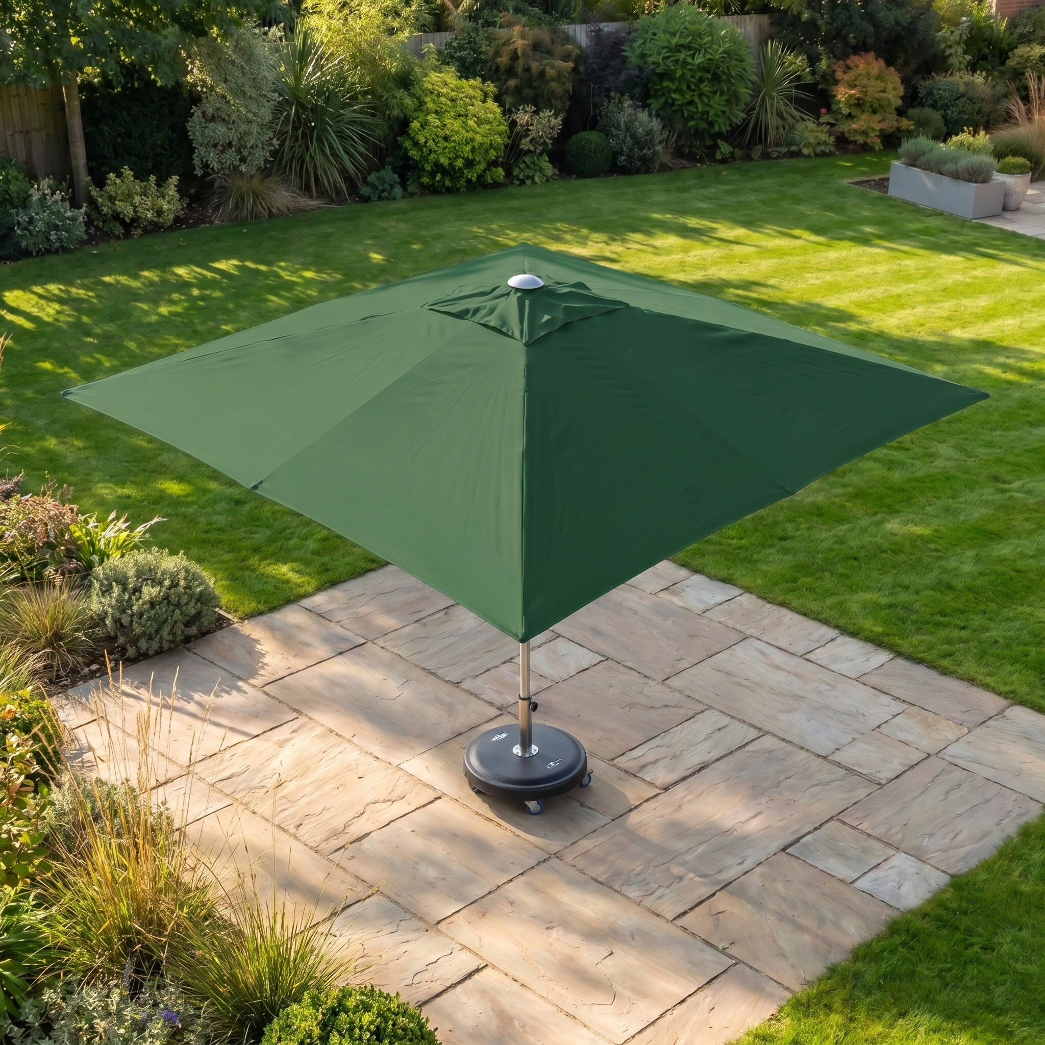 The Zelos 2.5m Square Parasol with LED Lighting in Green stands on a stone patio in a sunny, well-kept garden, providing ample shade and UV protection.