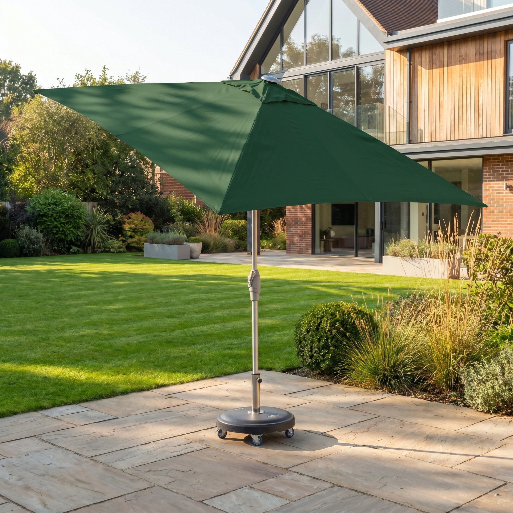 The Zelos 2.5m Square Parasol with LED Lighting in Green provides stylish shade and UV protection on a stone terrace in a modern, sunny garden outside the house.
