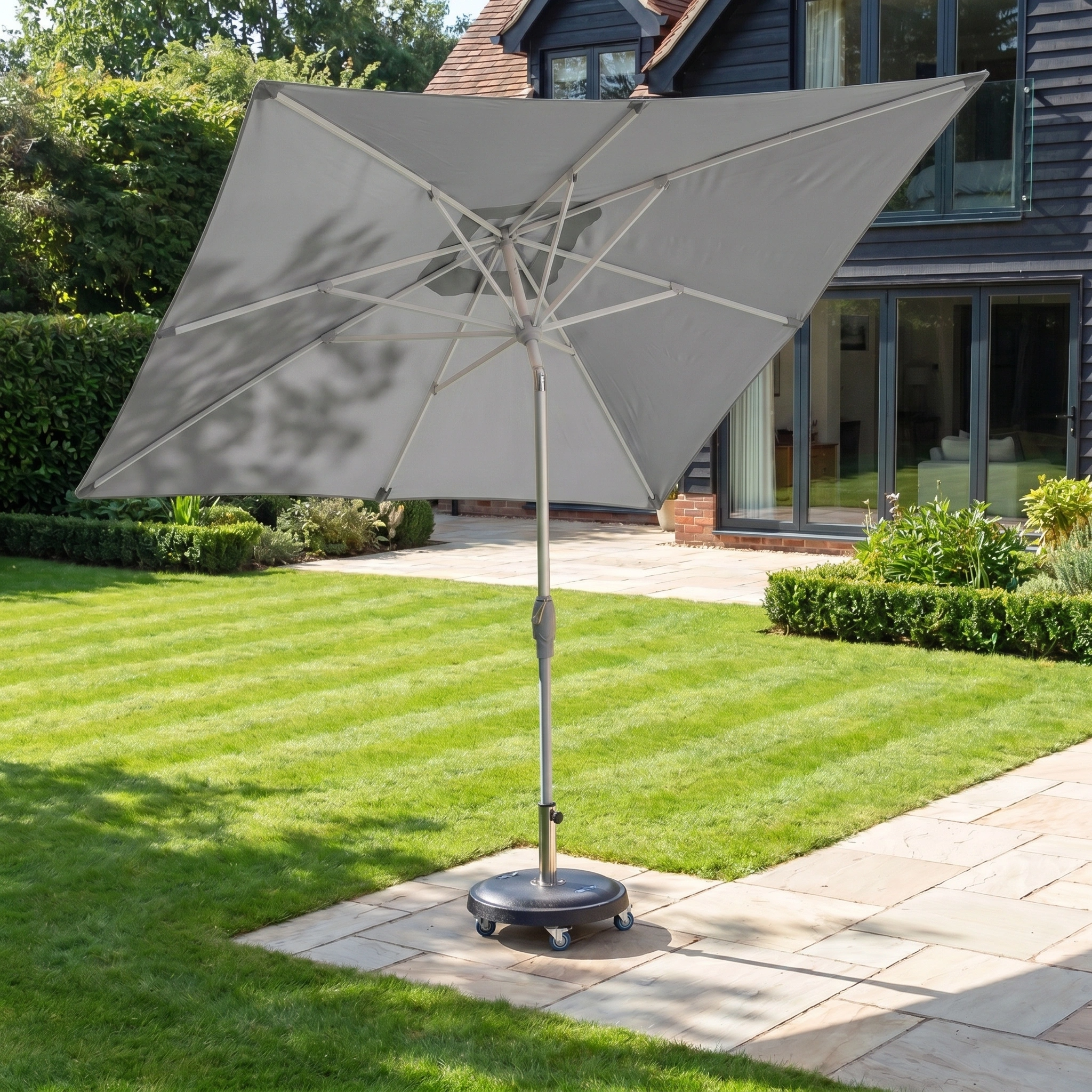 The Zelos 2.5m Square Parasol with LED Lighting in Grey stands open on a sunny, paved backyard, blending style and UV protection for outdoor comfort.