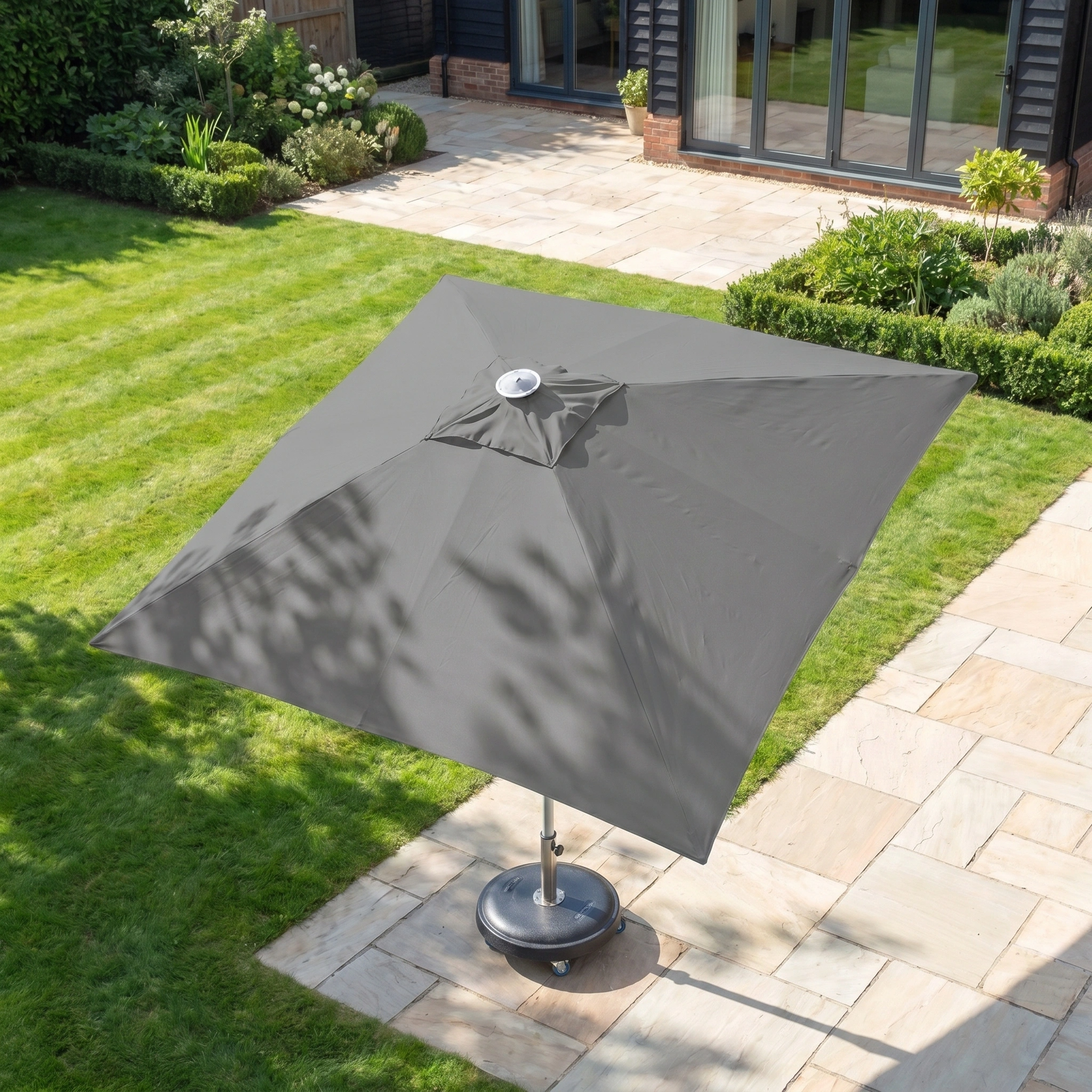 The Zelos 2.5m Square Parasol with LED Lighting in Grey provides shade and UV protection on a tiled patio beside a lush green lawn and garden, as viewed from above.