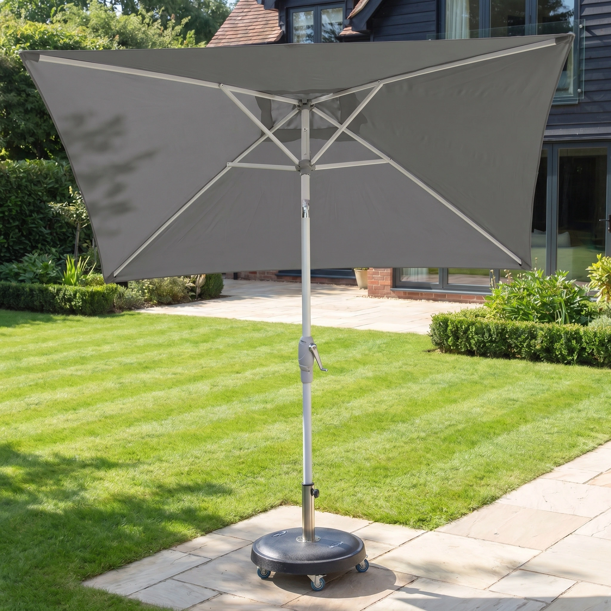 The Zelos 2.5m Square Parasol with LED Lighting in Grey stands on a wheeled base in a sunny, landscaped garden, providing ample UV protection.