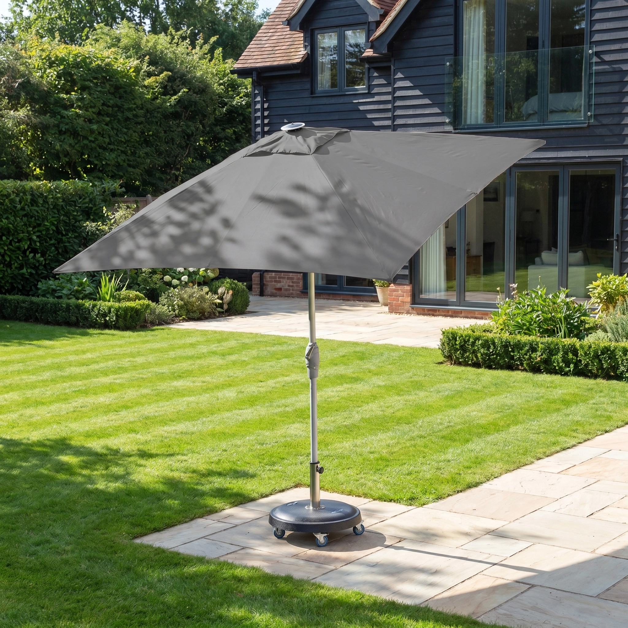The Zelos 2.5m Square Parasol with LED Lighting in Grey stands on a wheeled base on a paved patio, providing ample shade beside the green lawn.