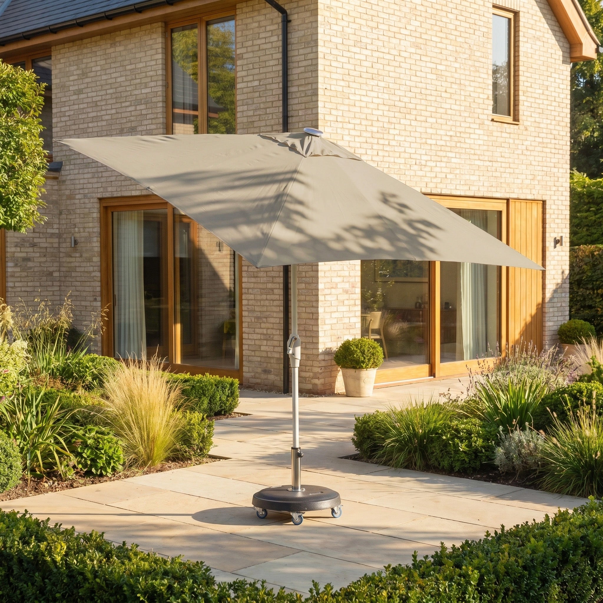 The Zelos 2.5m Square Parasol with LED Lighting in Taupe stands on a paved area in a landscaped garden outside a modern brick house, offering stylish shade and comfort.