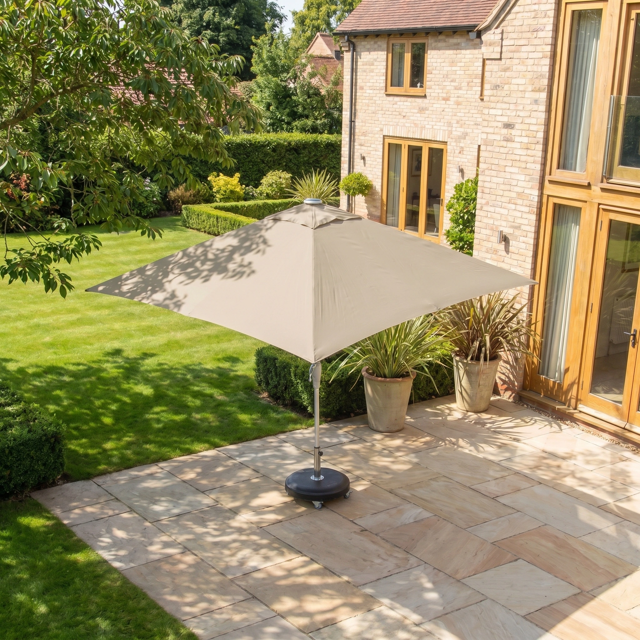 The Zelos 2.5m Square Parasol with LED Lighting in Taupe stands on a stone patio next to a brick house, surrounded by green lawn and plants, providing UV protection and stylish outdoor shade.