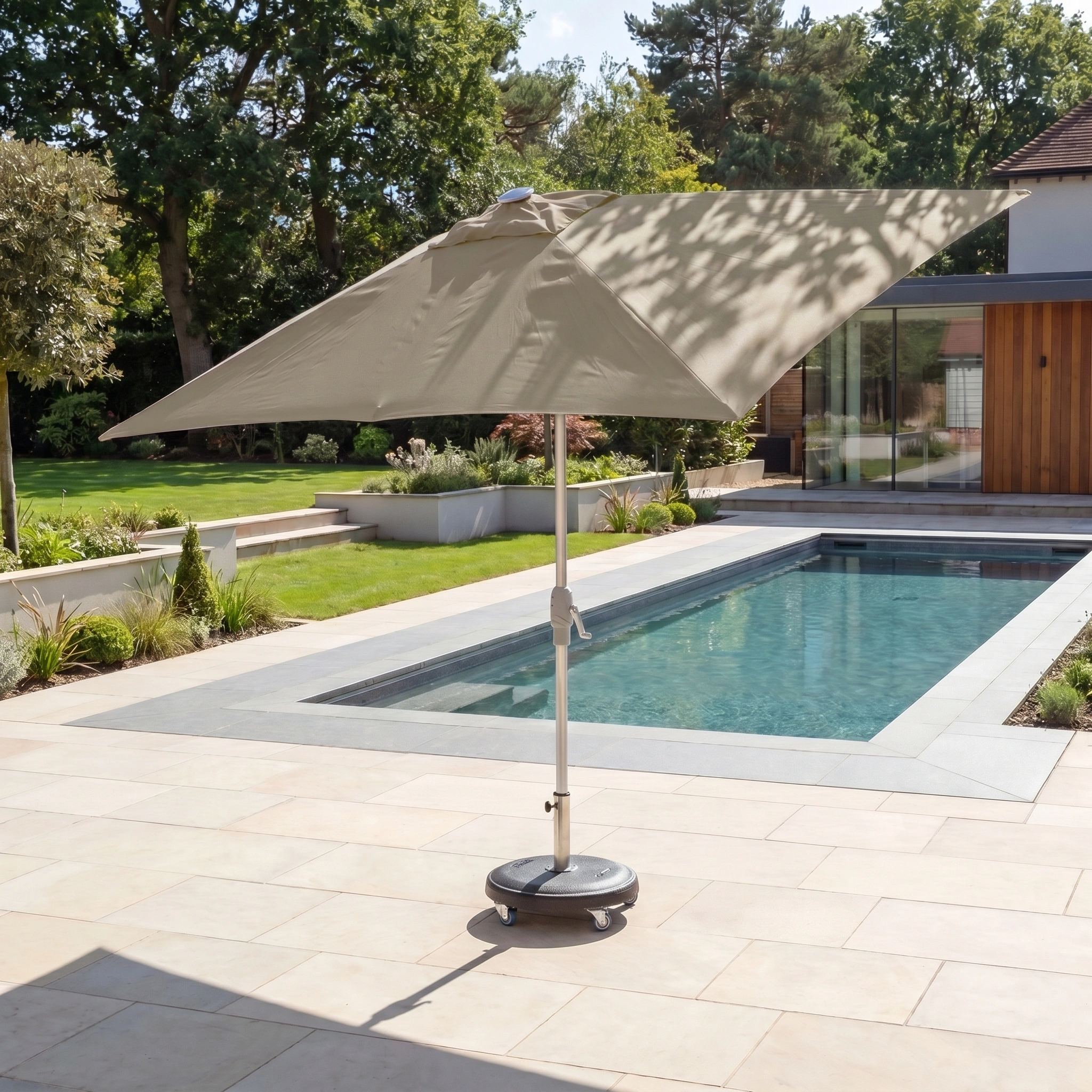 A Zelos 3m x 2m Rectangular Parasol with LED Lighting in Taupe stands beside a modern backyard swimming pool surrounded by greenery.