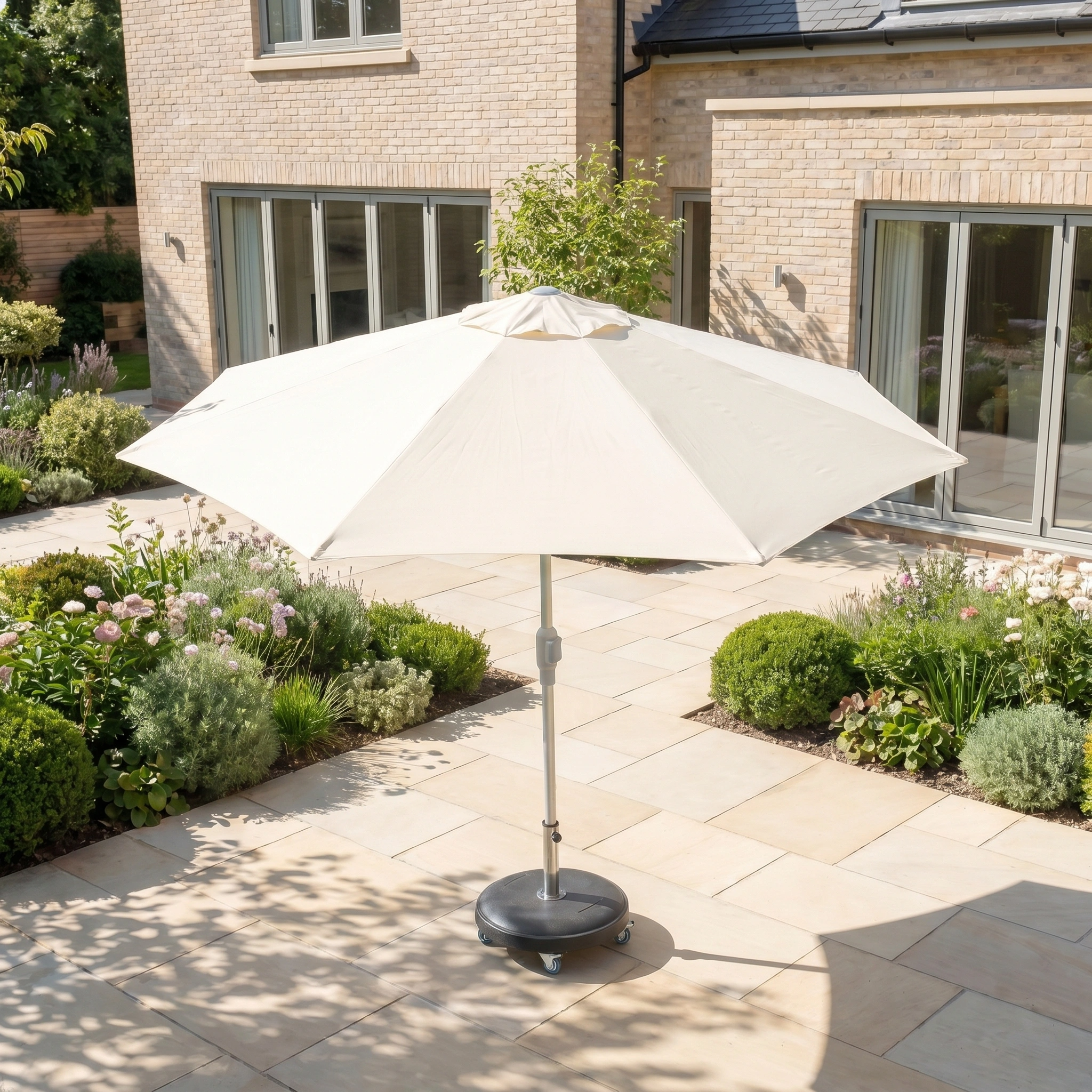 The Zelos 3m Round Parasol with LED Lighting in Cream stands open on a stone patio in a landscaped garden, offering stylish shade and UV protection.
