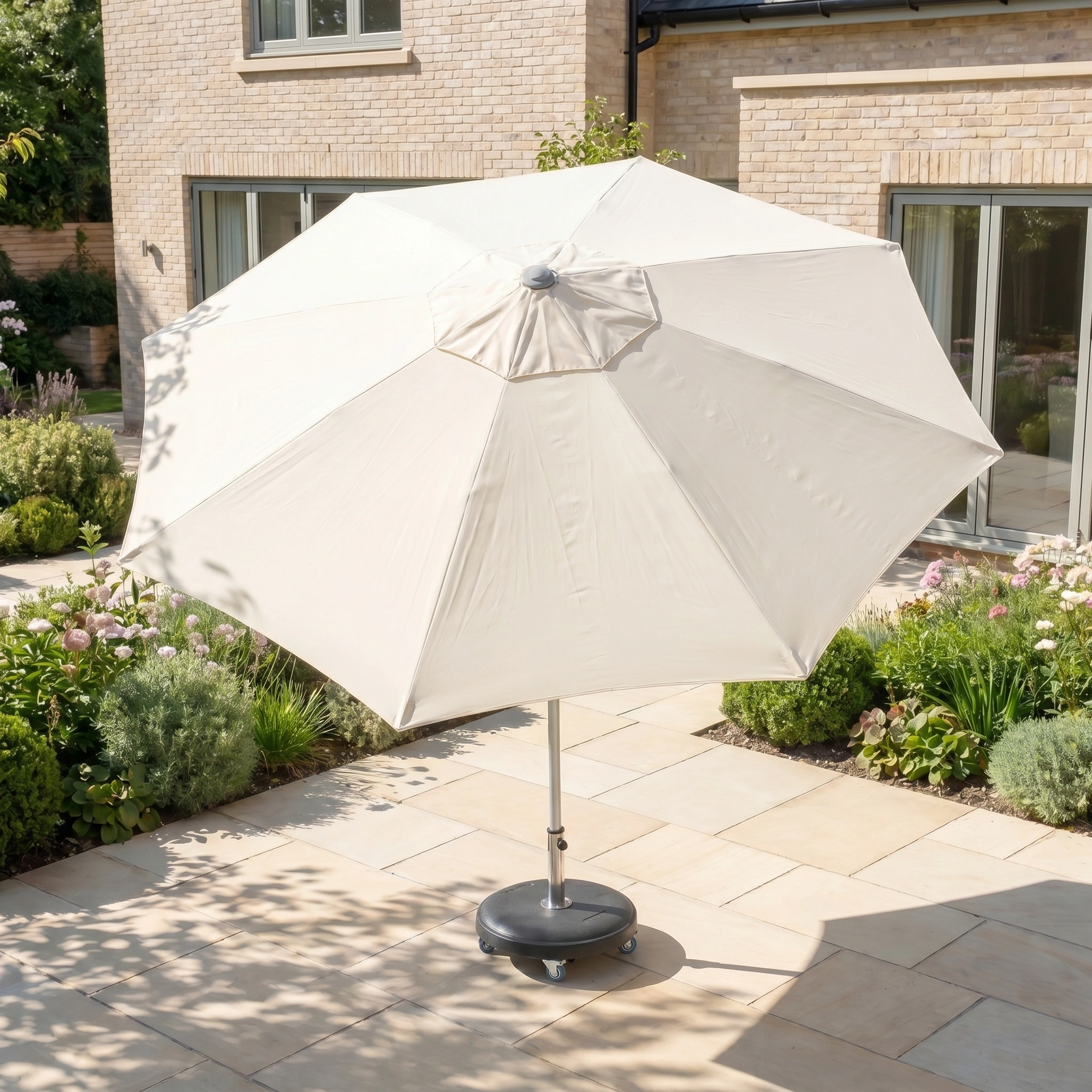 The Zelos 3m Round Parasol with LED Lighting in Cream stands on a tiled terrace in a sunny, landscaped garden, providing ample shade and UV protection.