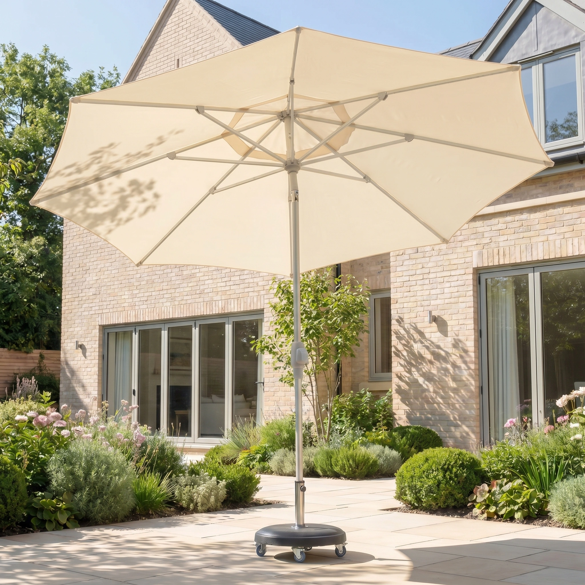 The Zelos 3m Round Parasol with LED Lighting in Cream shades a sunny stone terrace outside a modern brick house surrounded by garden plants.