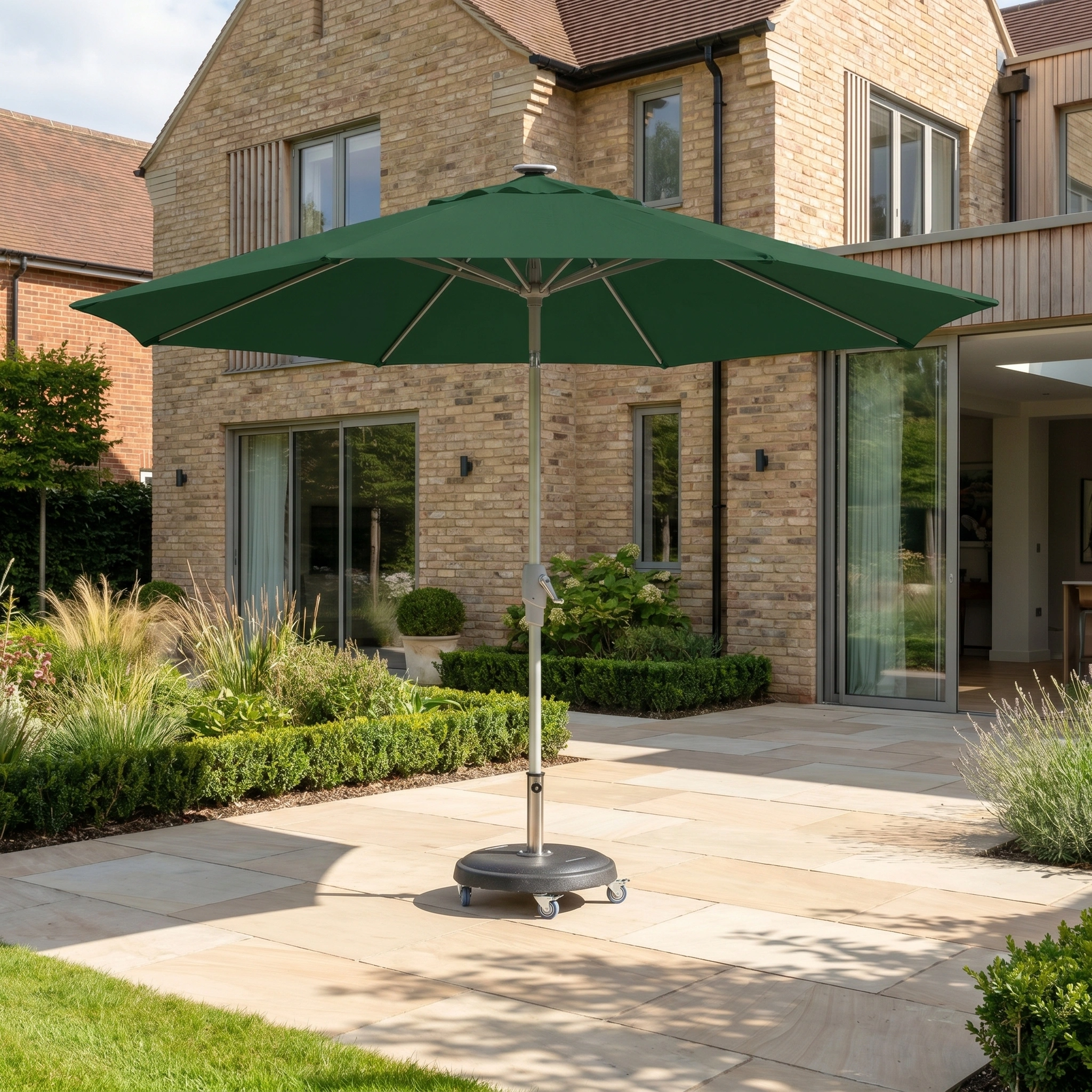 The Zelos 3m Round Parasol with LED Lighting in Green provides ample shade and UV protection on a modern terrace beside a brick house.