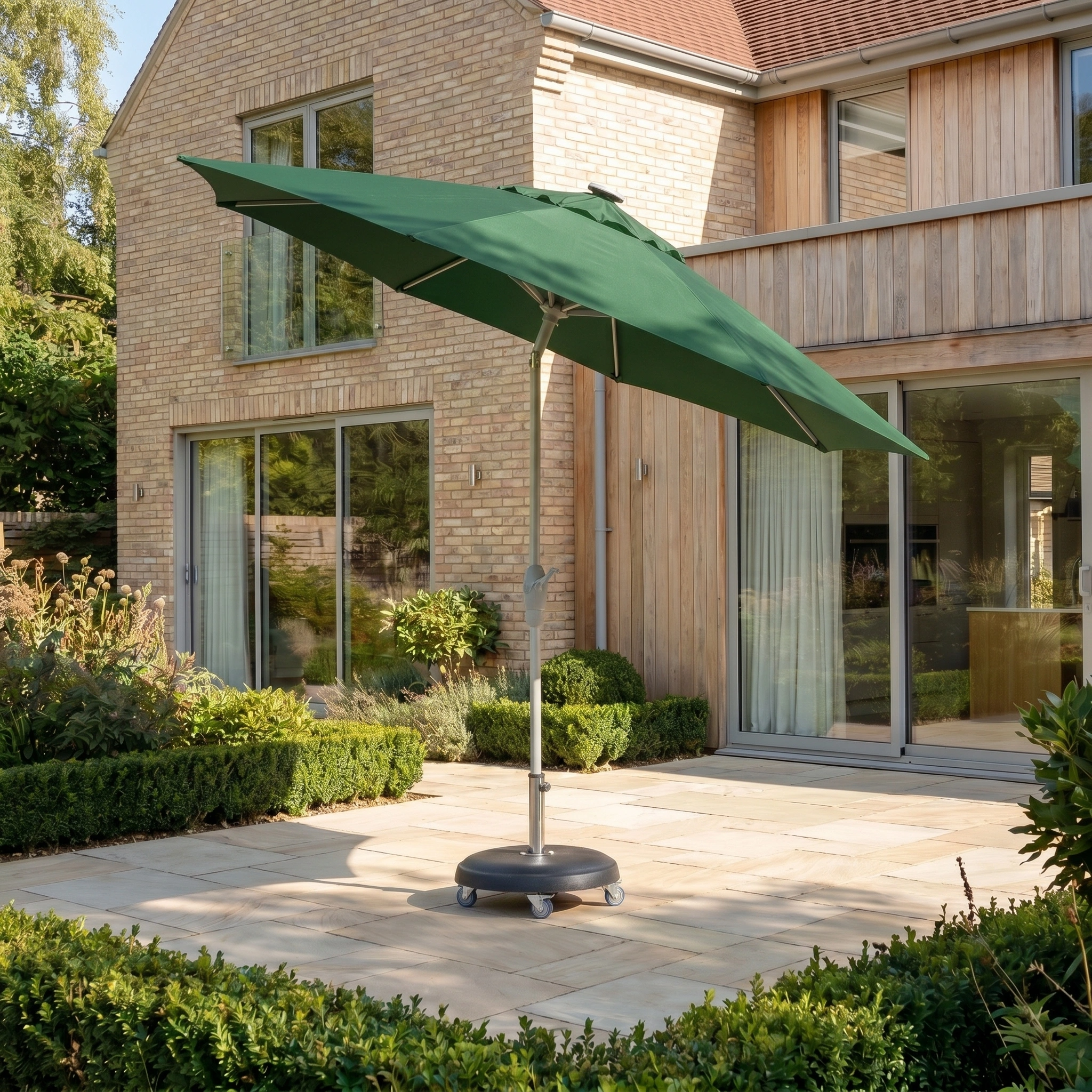 The Zelos 3m Round Parasol with LED Lighting in Green stands on a patio beside a modern house, providing UV protection and stylish shade.