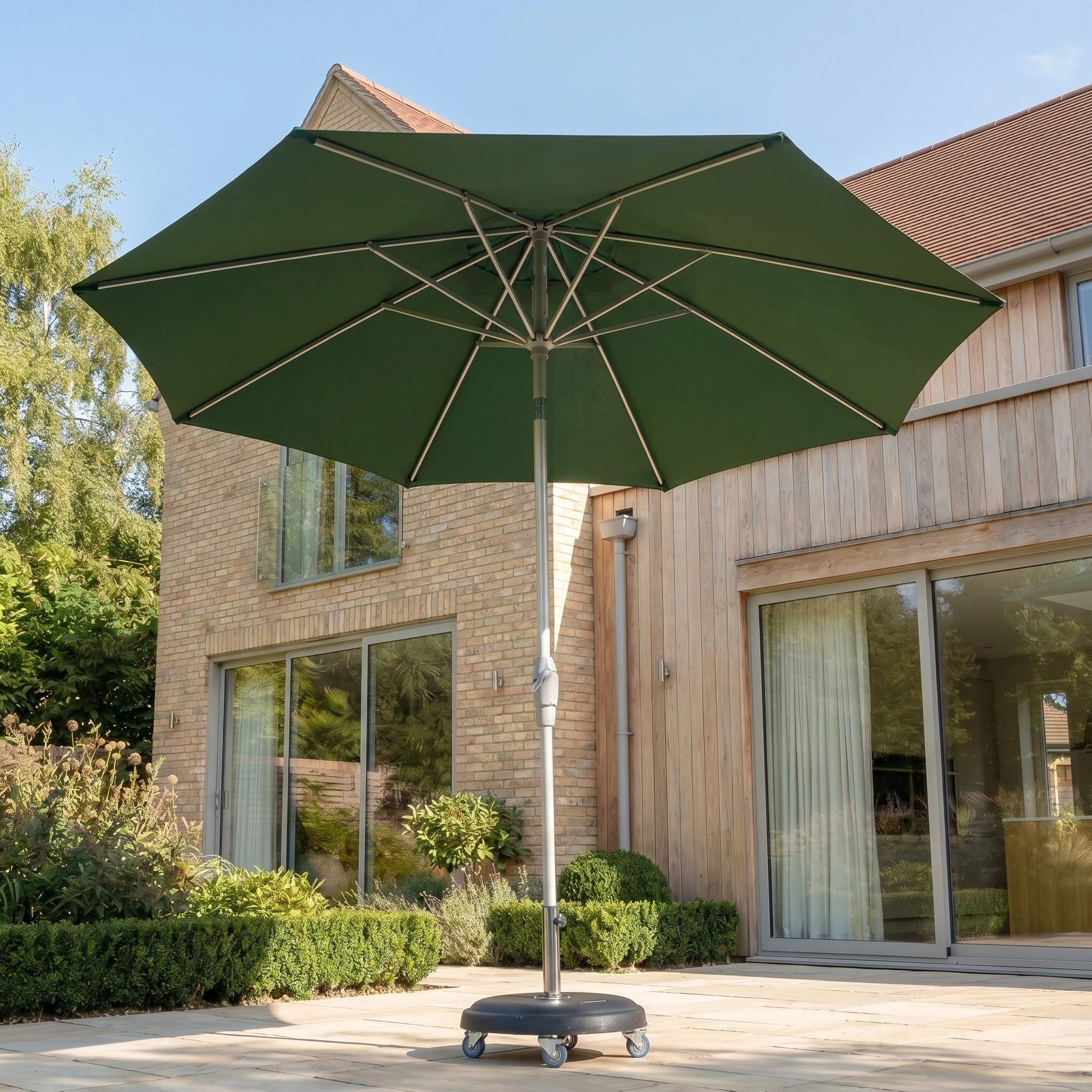 The Zelos 3m Round Parasol with LED Lighting in Green stands open on a paved area outside a modern brick house, providing UV protection to the patio below.