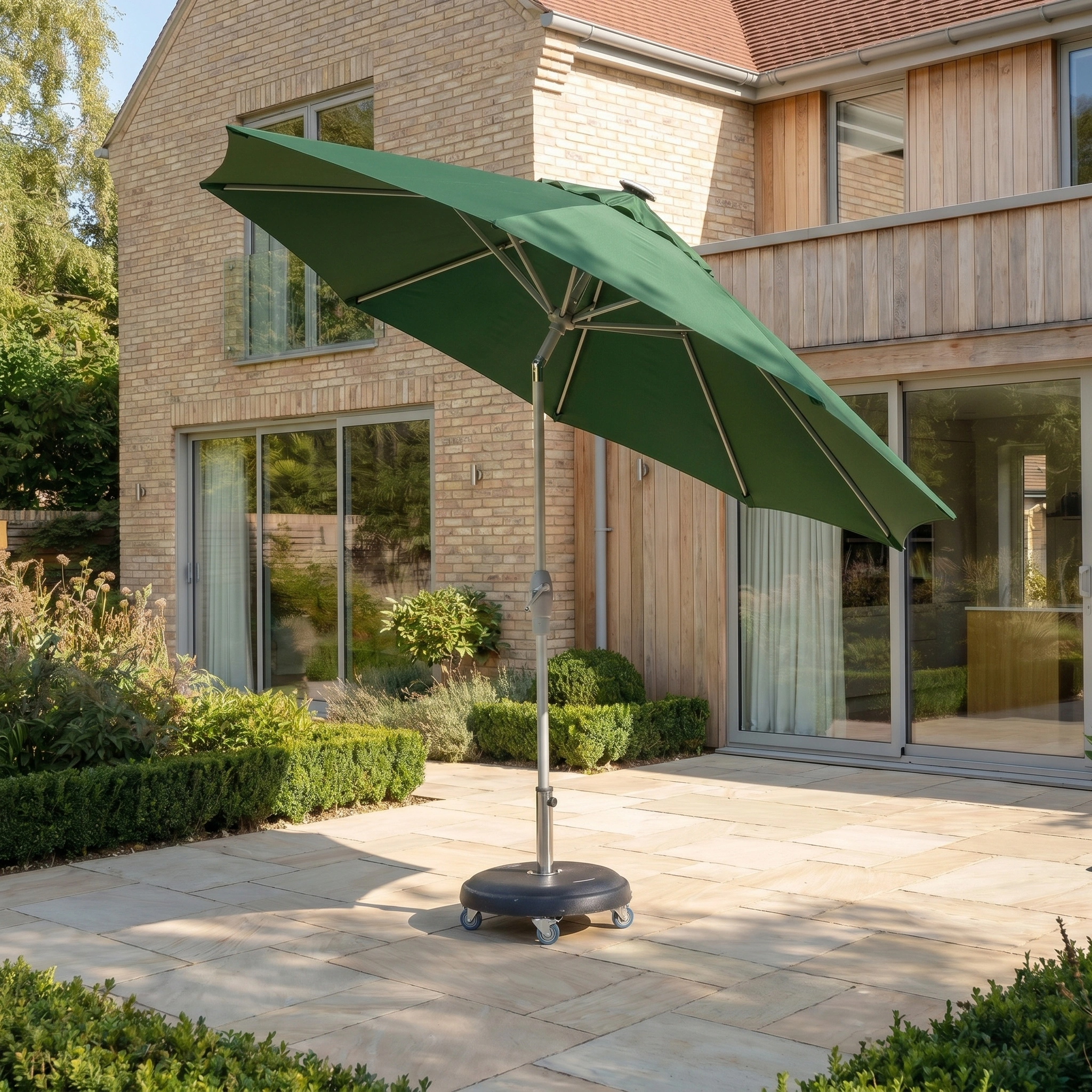 The Zelos 3m Round Parasol with LED Lighting in Green offers generous UV protection as it stands on a stone terrace outside a modern brick and wood house.