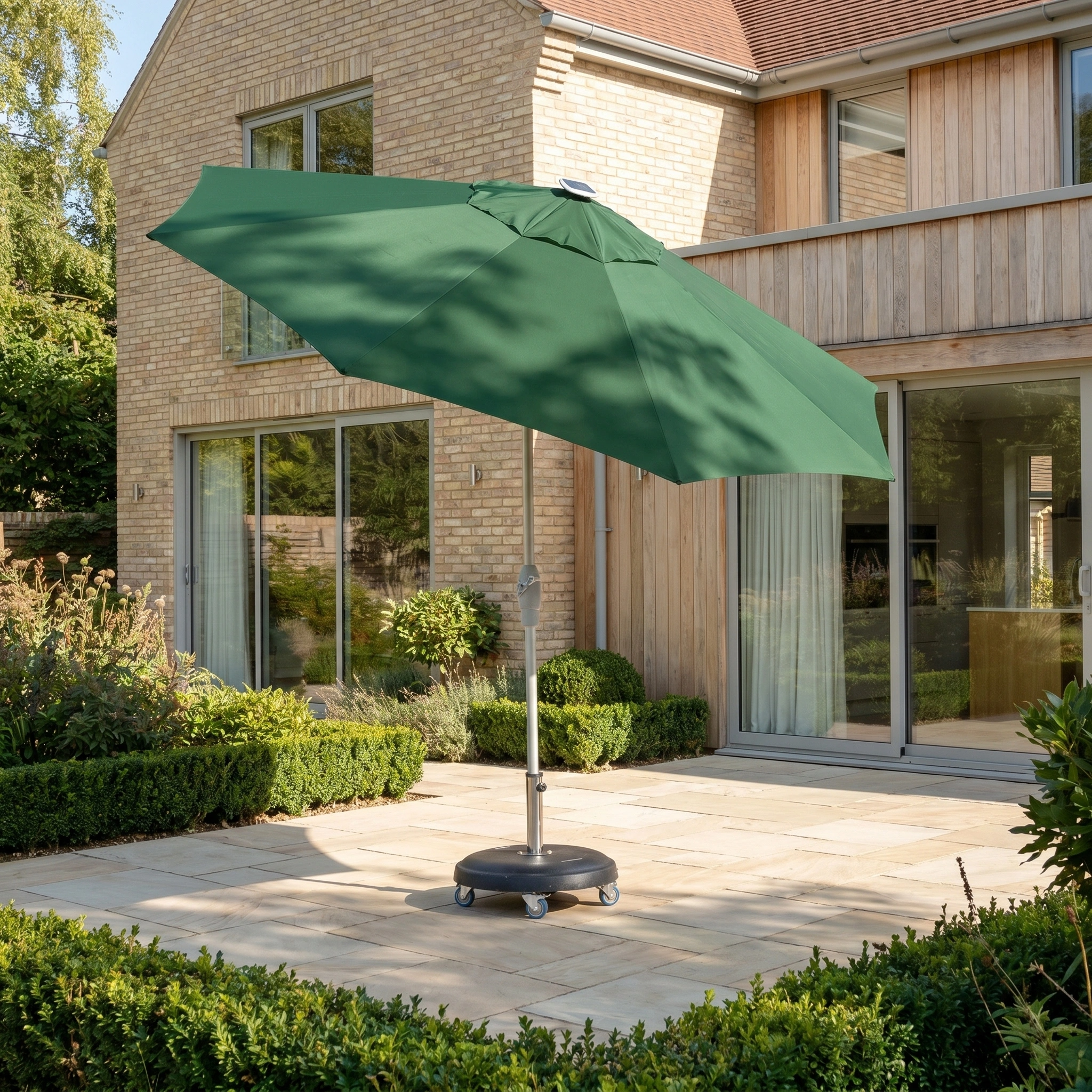 The Zelos 3m Round Parasol with LED Lighting in Green is set up on a stone terrace outside a modern house with large windows.