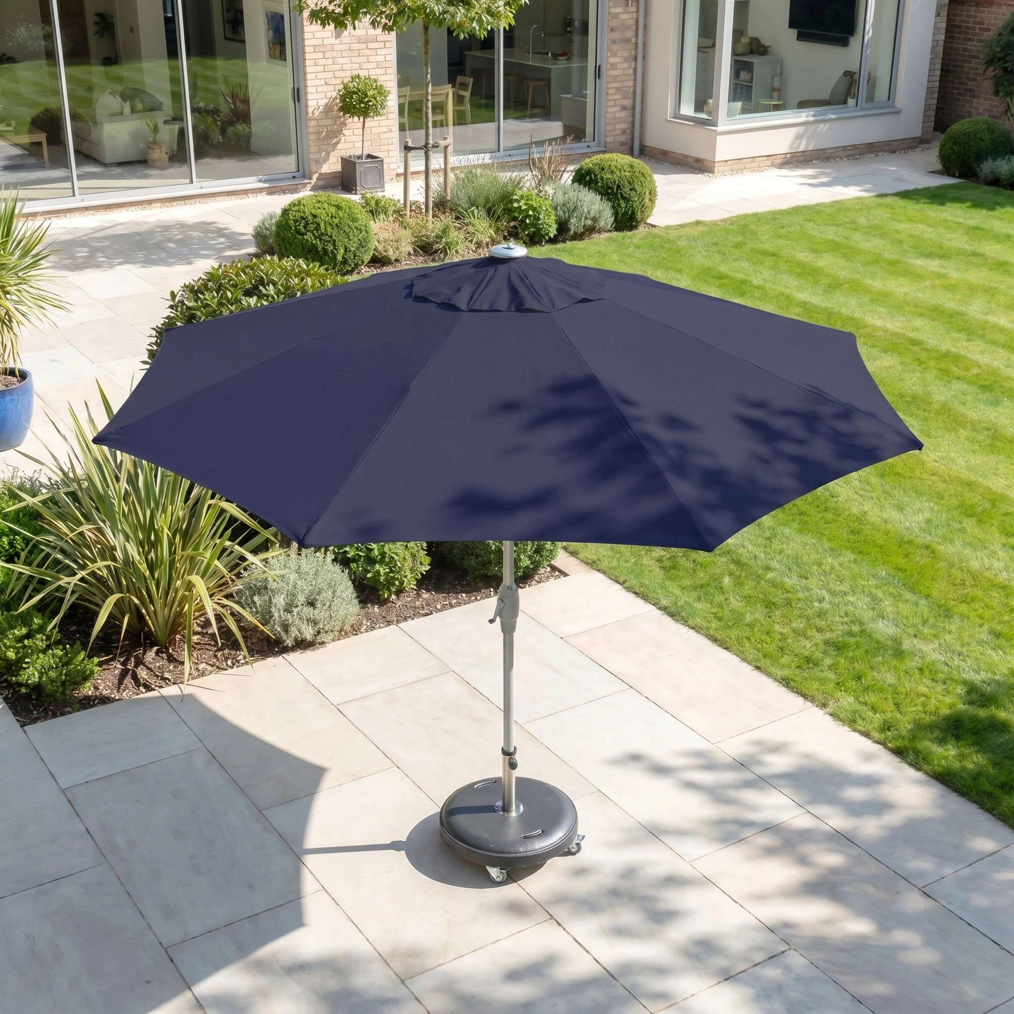 The Zelos 3m Round Parasol with LED Lighting in Navy stands on a tiled terrace beside a green lawn and bushes, offering stylish shade and UV protection.
