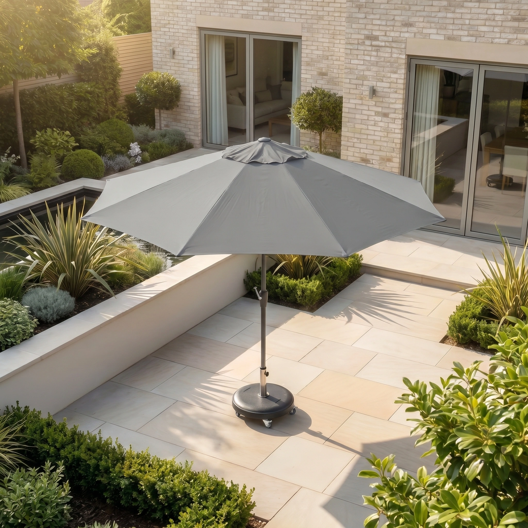 The Zelos 3m Round Parasol with LED Lighting in Grey stands in a sunny, modern garden with stone tiles and lush greenery, providing ample UV protection.