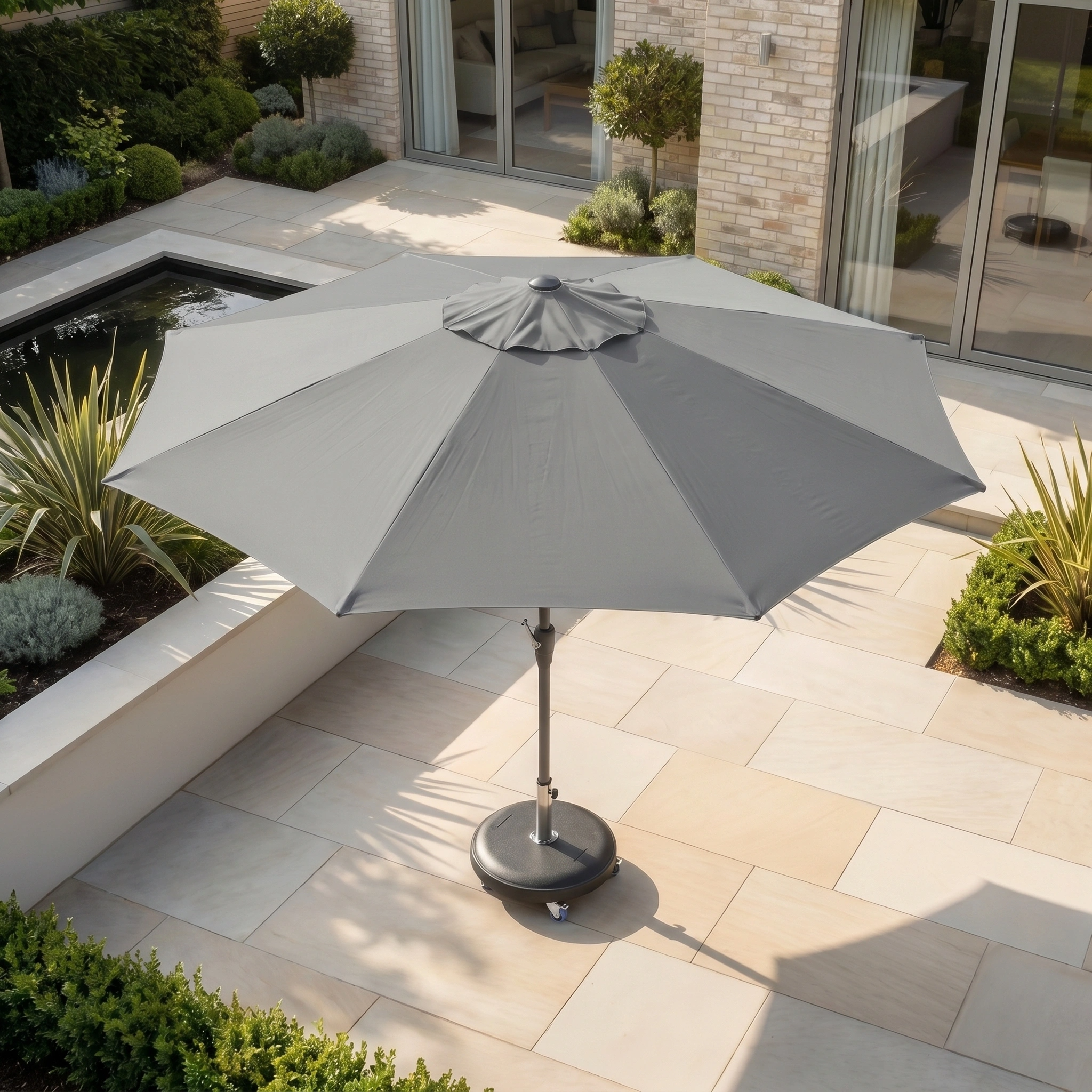 The Zelos 3m Round Parasol with LED Lighting in Grey stands open on a tiled terrace beside a small pond and garden, providing stylish shade and UV protection.