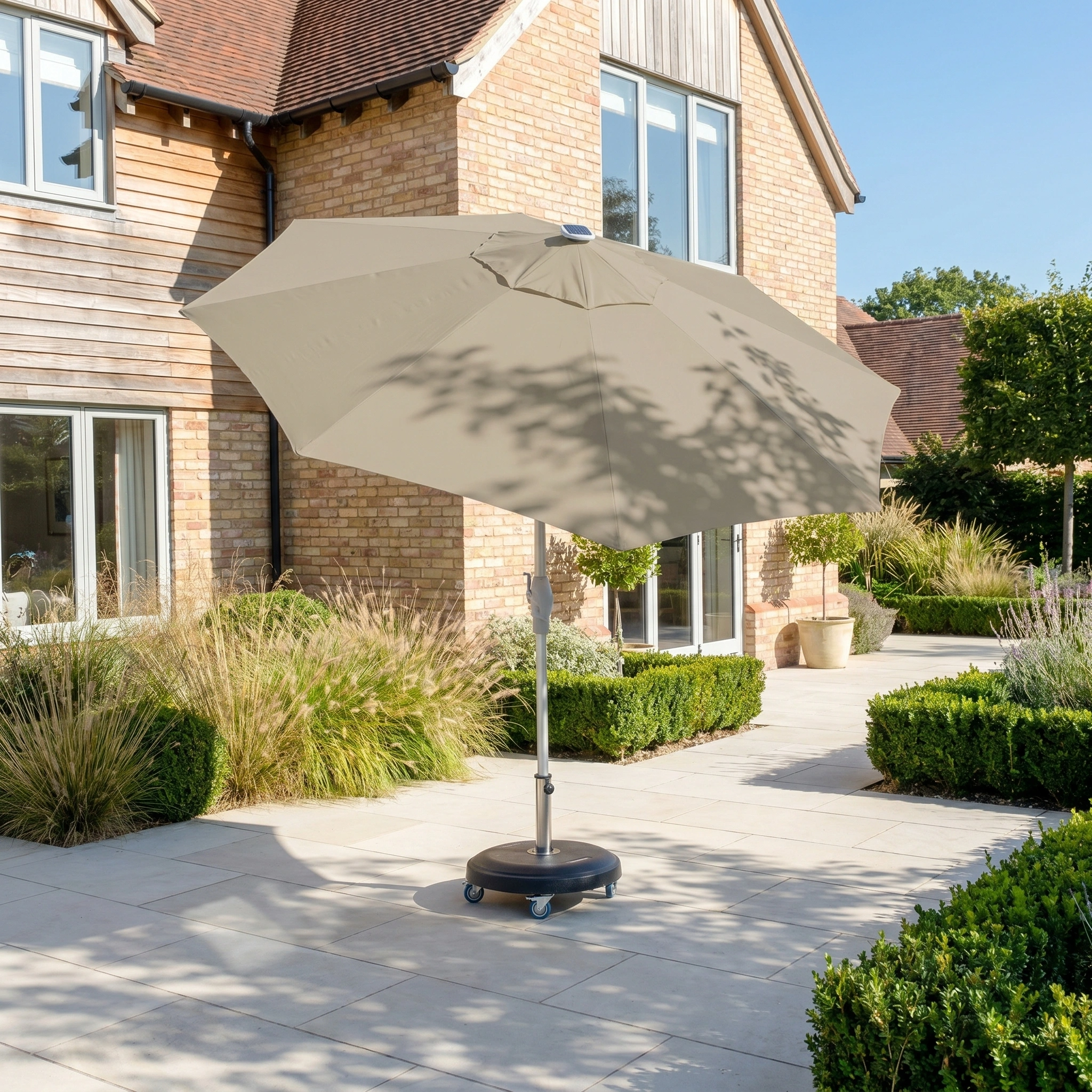 The Zelos 3m Round Parasol with LED Lighting in Taupe stands on a paved patio beside a brick house, providing stylish shade and ambient light.