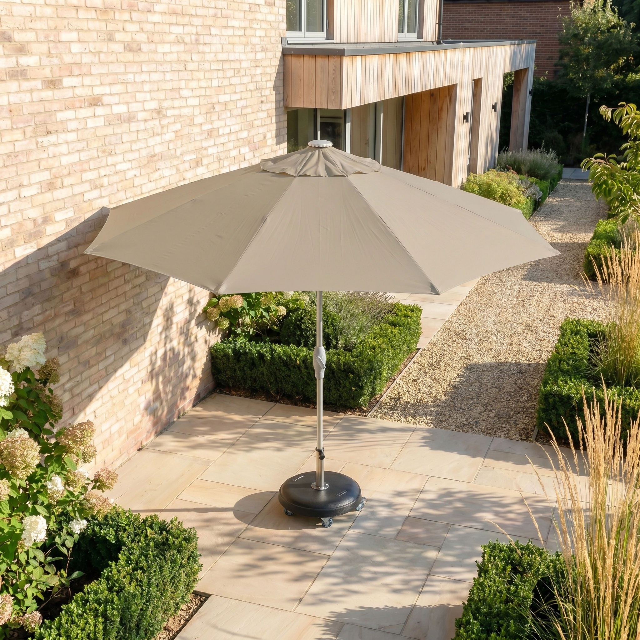 The Zelos 3m Round Parasol with LED Lighting in Taupe stands on a stone patio before a brick building, providing UV protection and cozy illumination for relaxing outdoor moments.