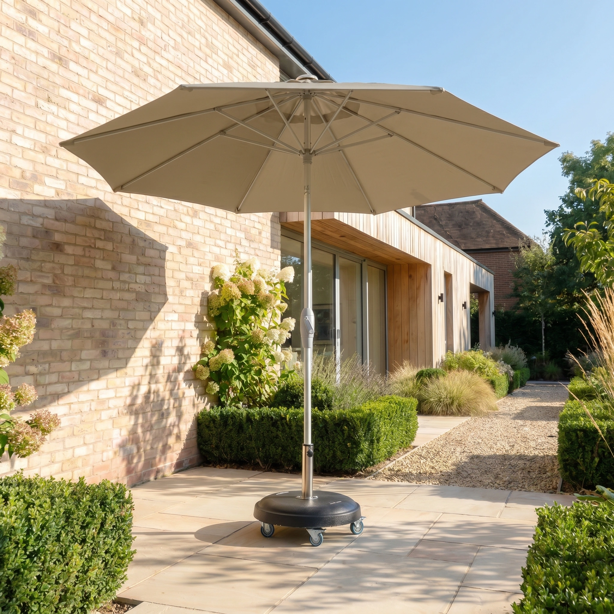 The Zelos 3m Round Parasol with LED Lighting in taupe stands on a stone patio beside a modern brick house, offering ample shade and UV protection.