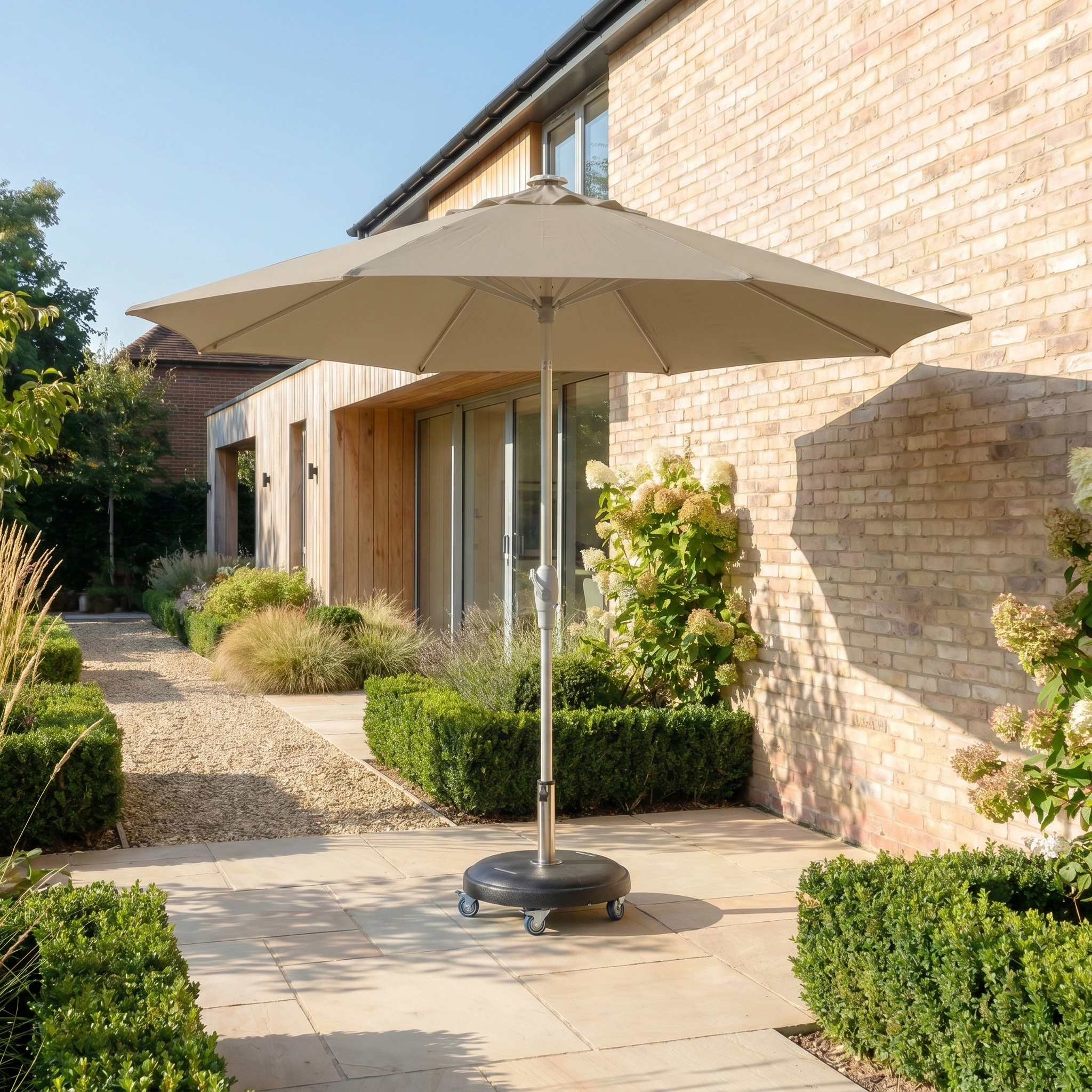 The Zelos 3m Round Parasol with LED Lighting in Taupe offers stylish shade and UV protection beside a modern brick house, enhancing your garden path with its elegant design and built-in illumination.