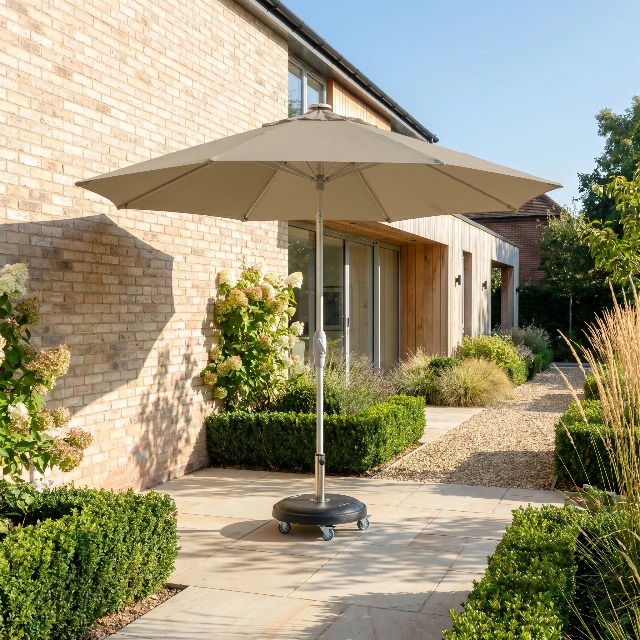 The Zelos 3m Round Parasol with LED Lighting in Taupe provides shade and style on a sunny stone pathway next to a modern brick house surrounded by garden plants.