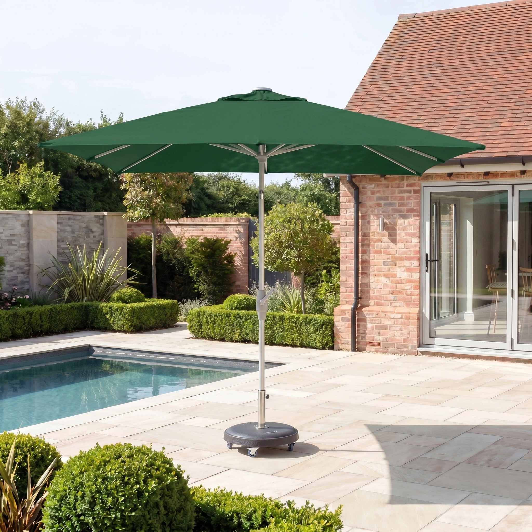 The Zelos 3m x 2m Rectangular Parasol with LED Lighting in Green stands by a swimming pool on a sunny stone-tiled patio near a brick house.