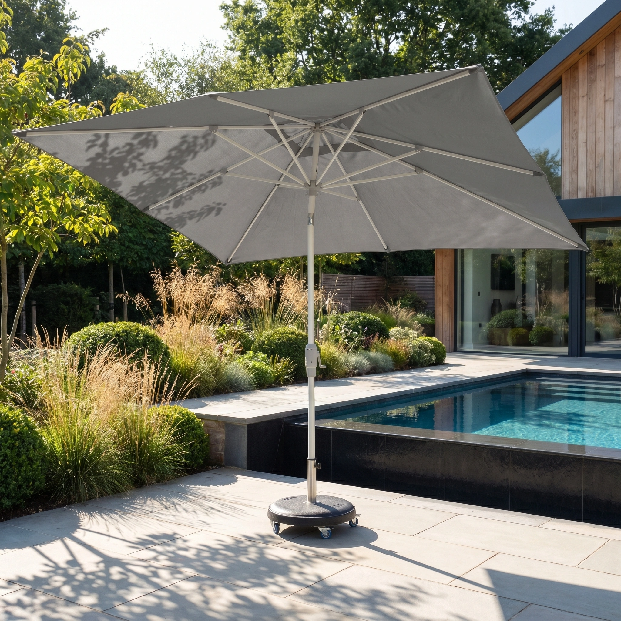The Zelos 3m x 2m Rectangular Parasol with LED Lighting in Grey stands beside a modern pool, surrounded by greenery and a wooden house.