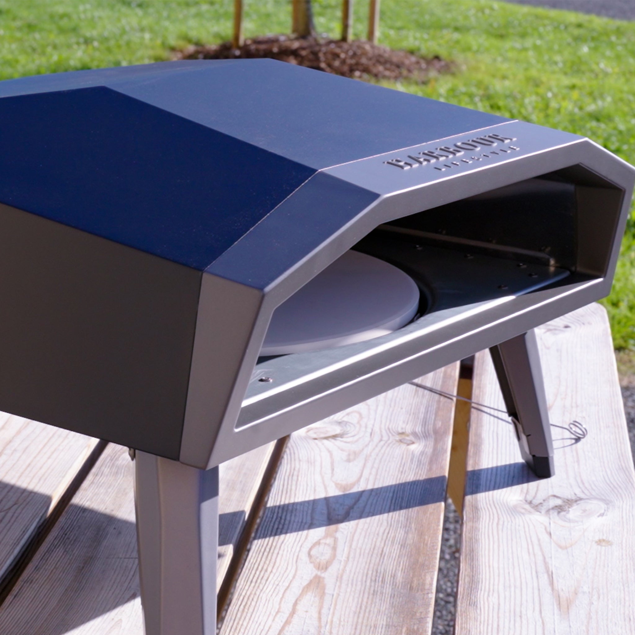 The Juno Pro 16" Gas Pizza Oven sits on a wooden table, ready to create a 60-second stone-baked pizza against a lush, grassy background.