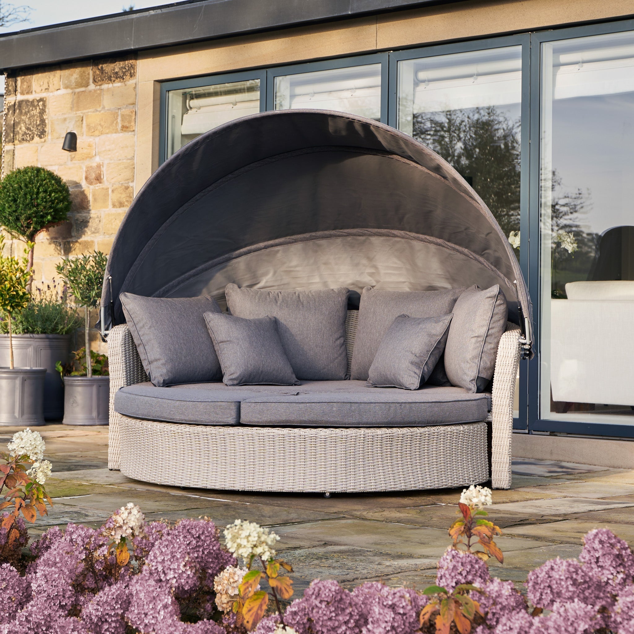 On the patio, the Pacific Lifestyle Bermuda Daybed in Stone Grey provides relaxation under its canopy amidst vibrant purple and white flowers. Made from weatherproof material, it elegantly complements a building with glass doors for a serene outdoor retreat.
