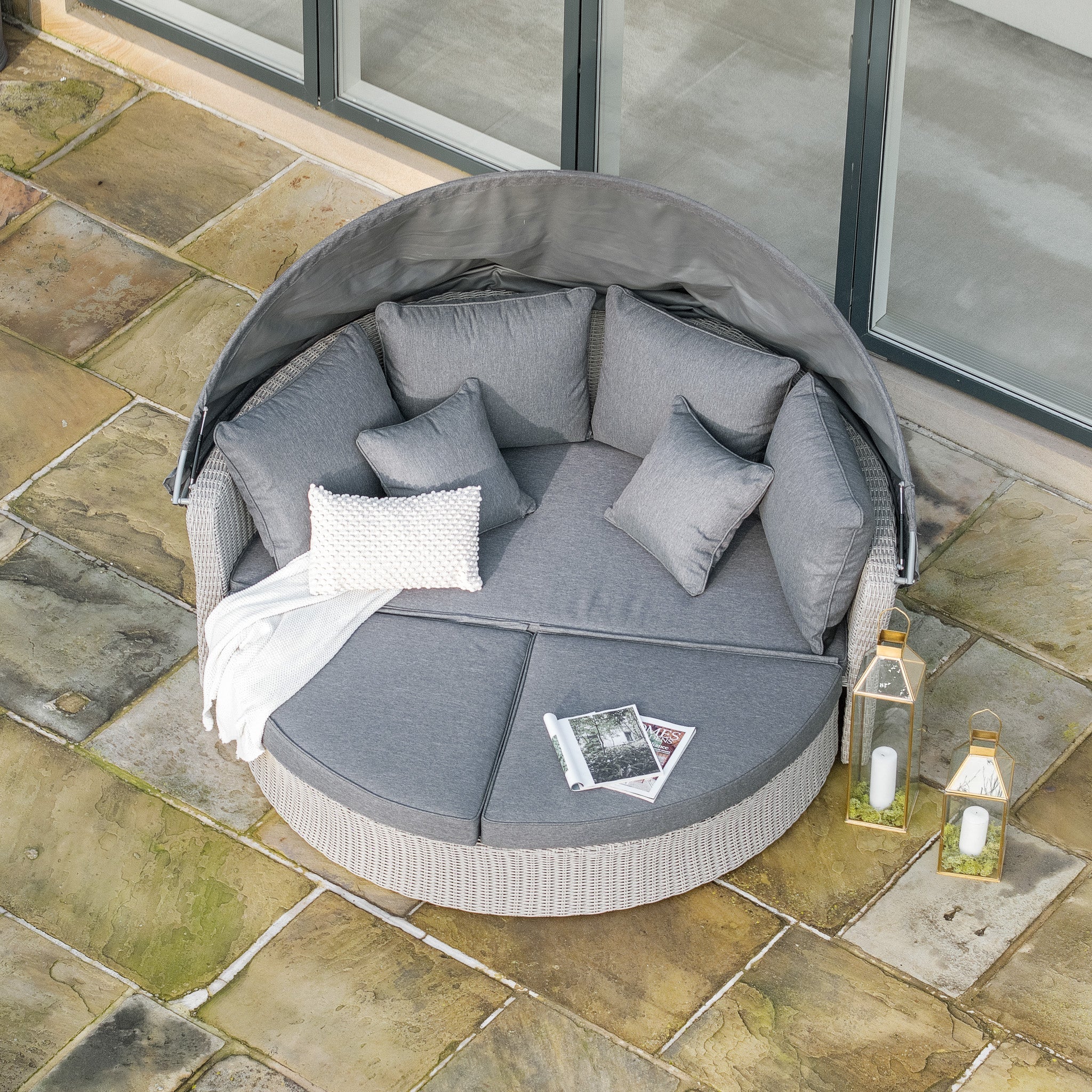 The Pacific Lifestyle Bermuda Daybed in Stone Grey, adorned with gray cushions and a knit throw, rests elegantly on the stone patio. Its weatherproof material ensures long-lasting use, while two lanterns provide a cozy ambiance.