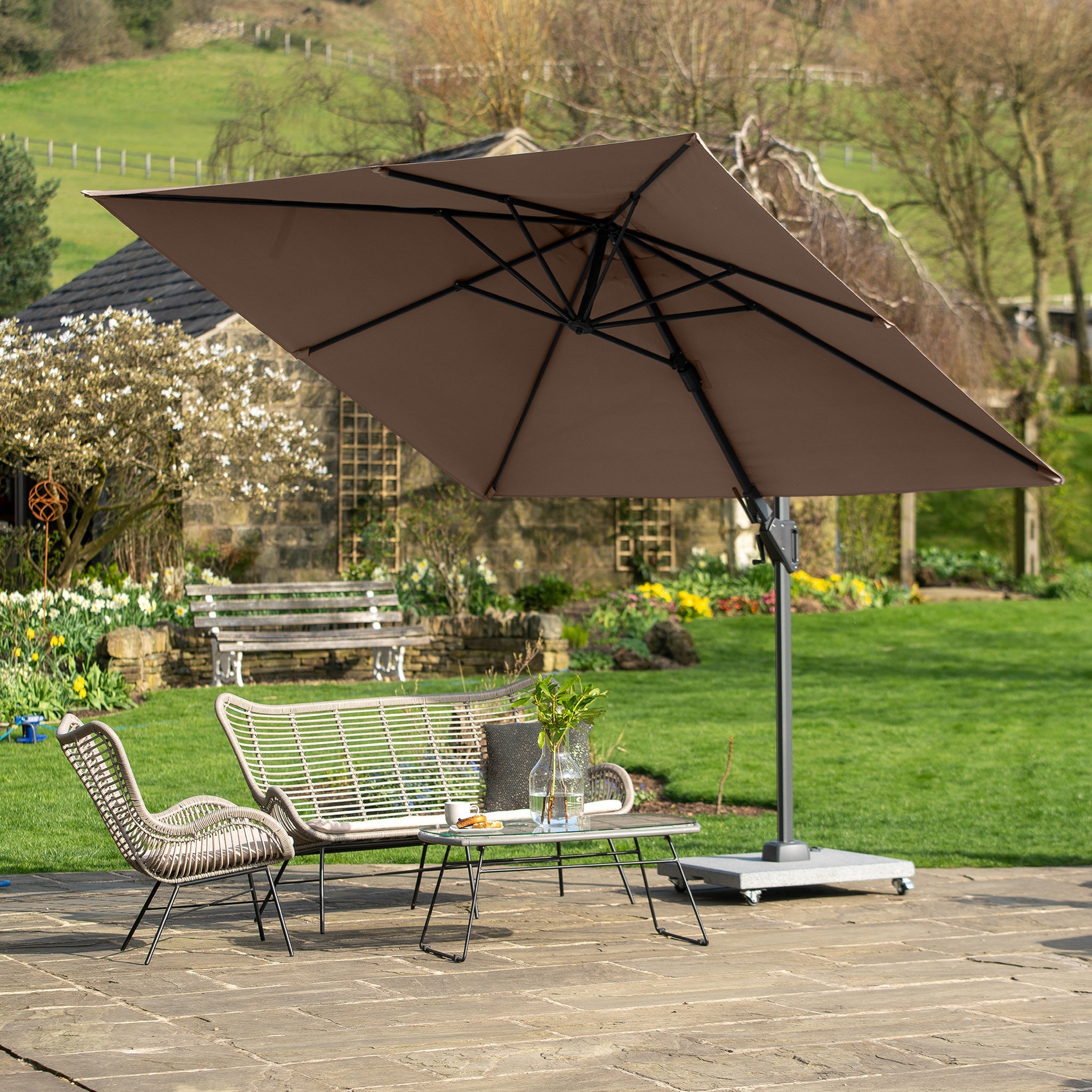 A patio features wicker chairs and a table on stone pavement with a lush garden view, shaded by a Pacific Lifestyle Voyager T2 2.7m Square Parasol in Grey.